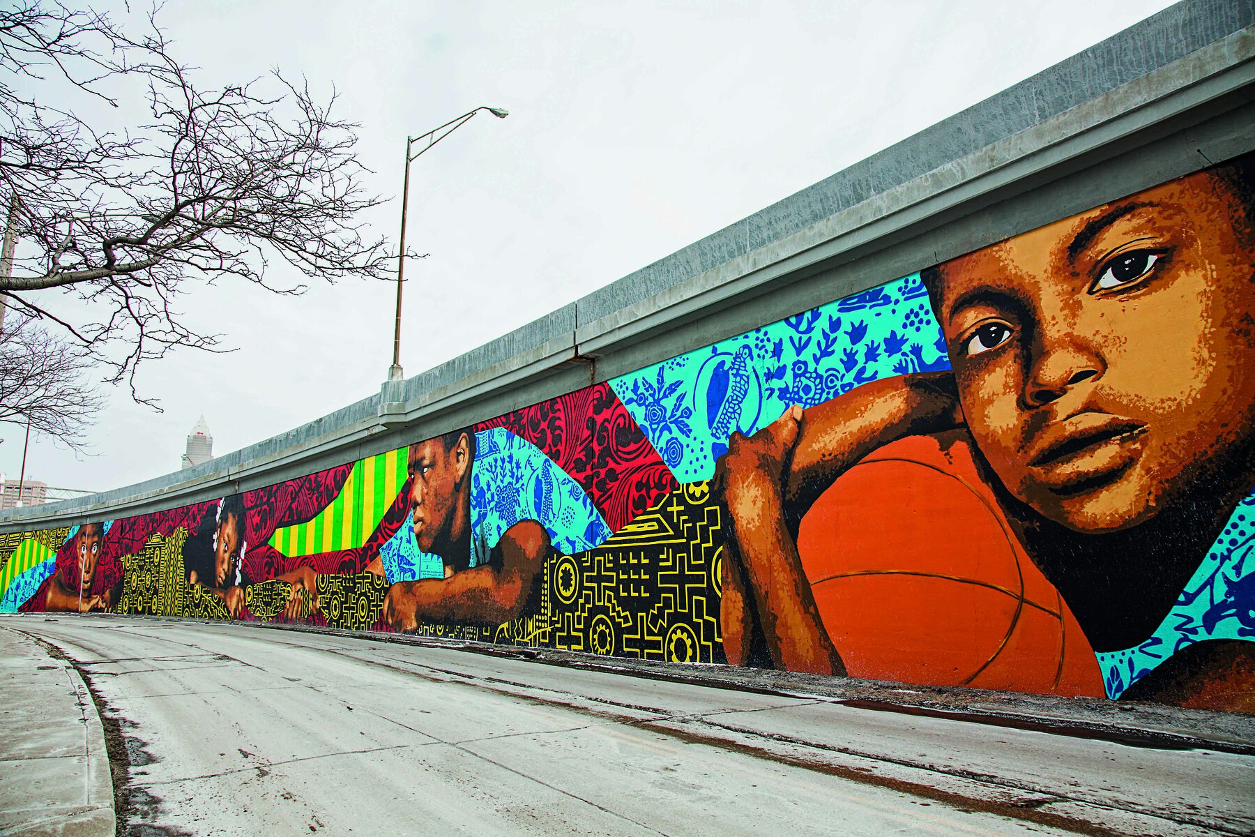 Fotografia. Vista de pintura mural em um viaduto, mostrando crianças e jovens como se estivessem debruçados sobre um muro. Todos são afrodescendentes. No primeiro plano, há um menino de cabelo curto preto, segurando uma bola de basquete. Ao fundo e ao redor das figuras humanas há desenhos simétricos em preto e amarelo e outros elementos decorativos em verde e amarelo, em tons de azul e em vermelho e preto.