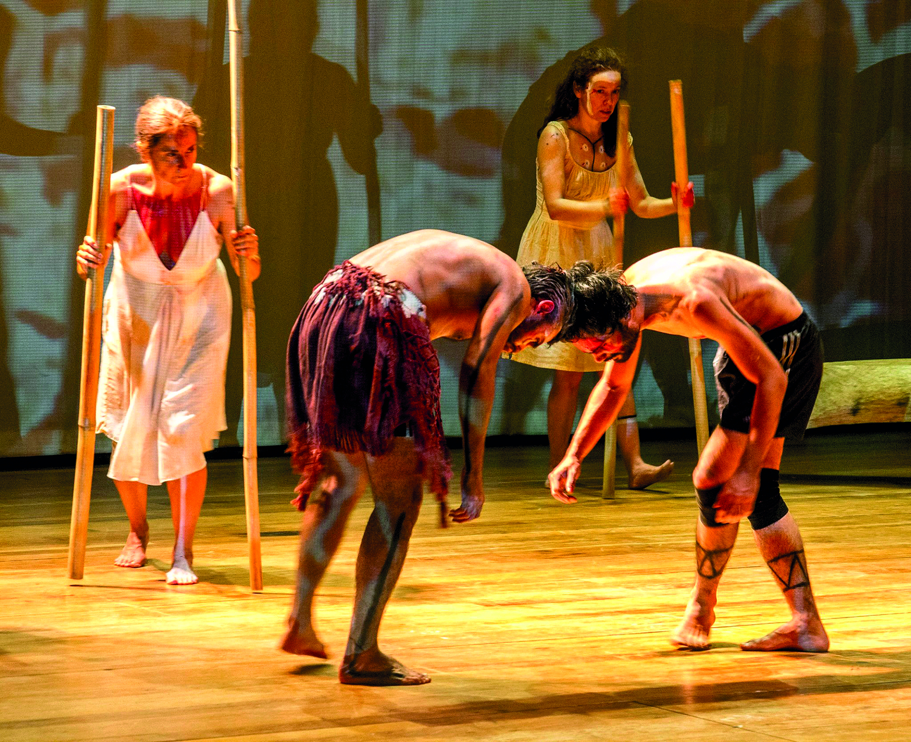 Fotografia. Cena teatral. No palco, há quatro atores, dois homens e duas mulheres. Os dois homens ocupam o centro da cena. Estão recurvados apoiando uma cabeça contra a outra. Ambos estão de peito nu, vestindo bermudas e descalços. O homem da esquerda traz, sobre a bermuda, uma saia curta, marrom-avermelhada, de plumas. O da direita, usa joelheiras pretas. Ambos apresentam pinturas corporais de linhas finas, em preto e cinza.  Mais ao fundo, à esquerda, há uma mulher de cabelo loiro preso e, à direita, uma mulher de cabelo escuro longo. Ambas usam vestido branco, estão descalças e têm pinturas corporais. Cada uma delas segura dois bastões, um em cada mão, que batem no chão enquanto se movimentam.