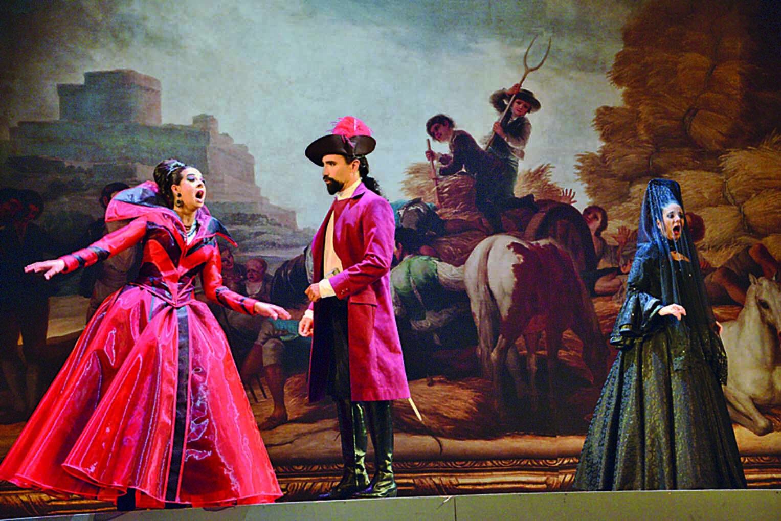 Fotografia. Cena de espetáculo de ópera. Em primeiro plano à esquerda, uma mulher de cabelo preto preso e vestido de época longo, vermelho, canta interagindo com um homem de chapéu de duas pontas preto adornado de vermelho trajando camisa branca de punhos largos, calça preta e sobretudo vermelho. Usa botas de cano longo, com enfeites prateados. À direita, quase de costas para o homem, há uma mulher de chapéu alto, em formato de cone, coberto por um véu azul preso ao topo, que cai e envolve seus ombros. Usa vestido verde-escuro e está cantando. O cenário é composto de uma pintura grande que recobre todo o fundo do palco. Na pintura, há cavalos pastando e cavaleiros conversando, Dois jovens montados em um animal que leva uma carga de feno empunham ferramentas. Um deles usa chapéu e, equilibrado sobre a montaria, segura um grande garfo de ferro apontado para cima. Ao fundo, à esquerda, sob uma luz difusa, há um grande castelo em formato retangular. À direita, há uma pilha enorme de fardos de feno. O ceú é azul-claro no centro e coberto de nuvens escuras nas laterais.