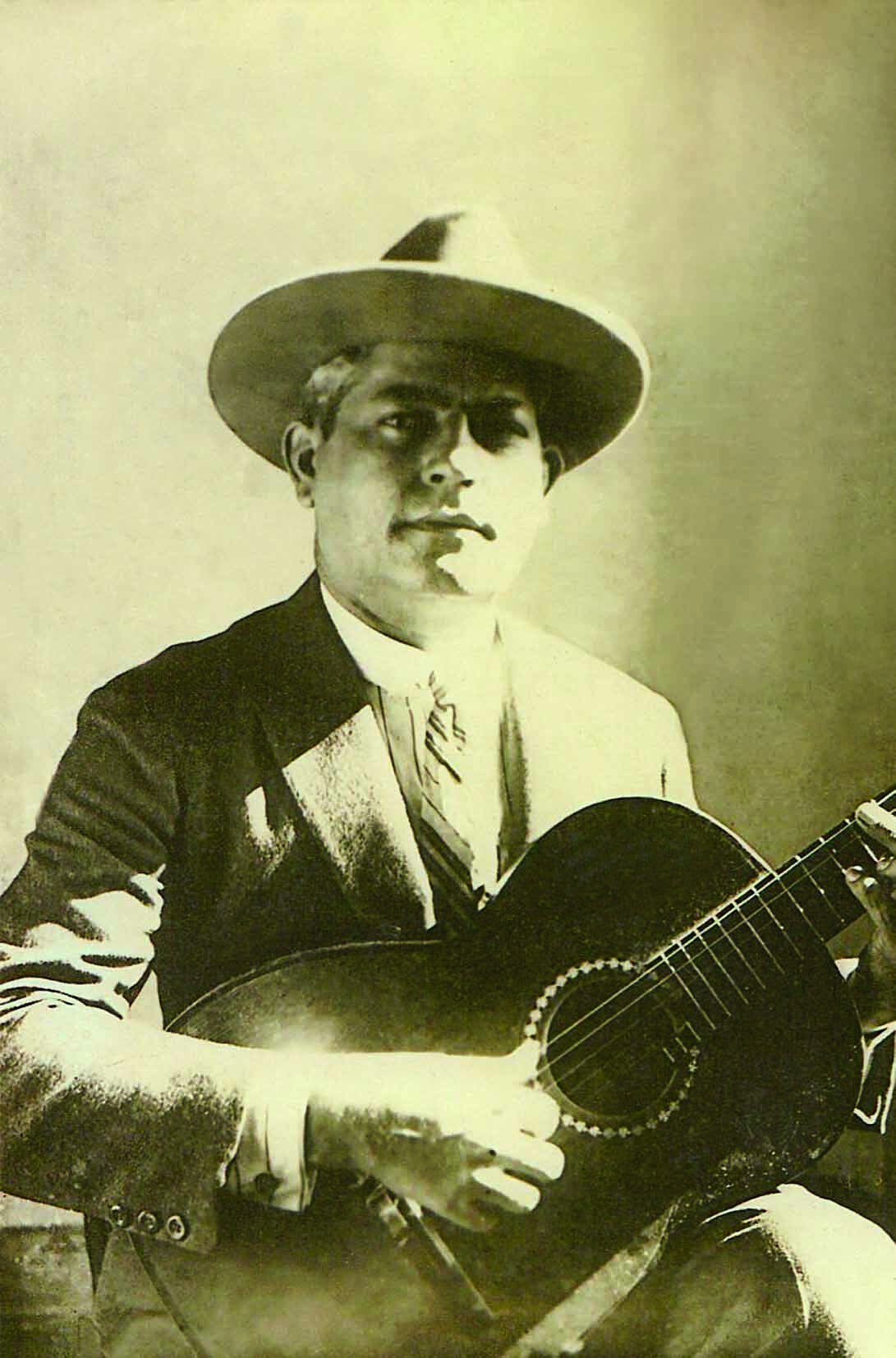Fotografia em preto e branco. Retrato de homem de cabelo curto e chapéu com aba redonda, vestindo camisa branca, coleta, paletó e gravata. Ele segura um violão em posição de tocá-lo.