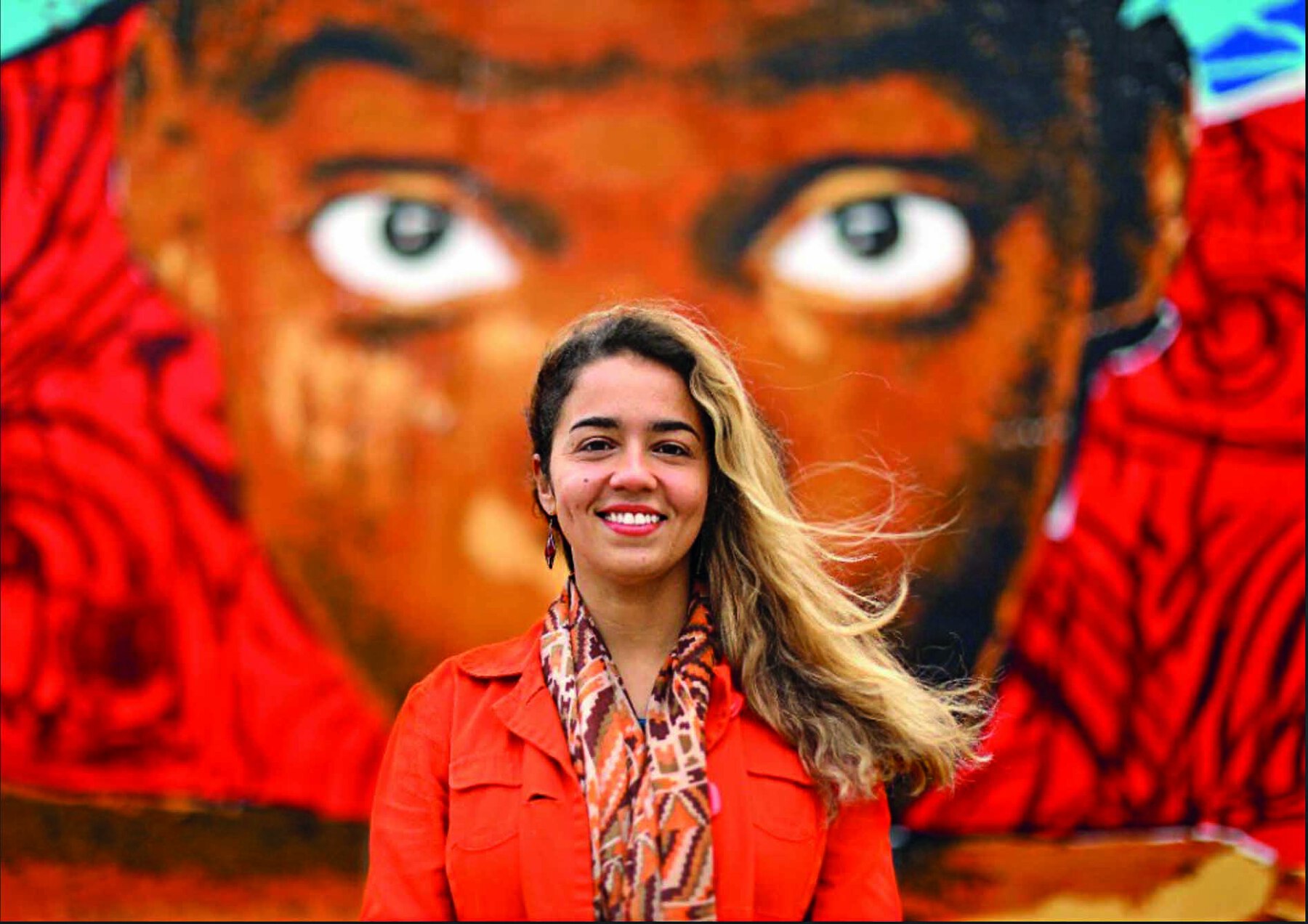 Fotografia. Mulher de cabelo longo loiro, vestindo blusa com estampas nas cores marrom e laranja e jaqueta laranja. Atrás dela há pintura desfocada do rosto de um menino destacando seus olhos castanhos.