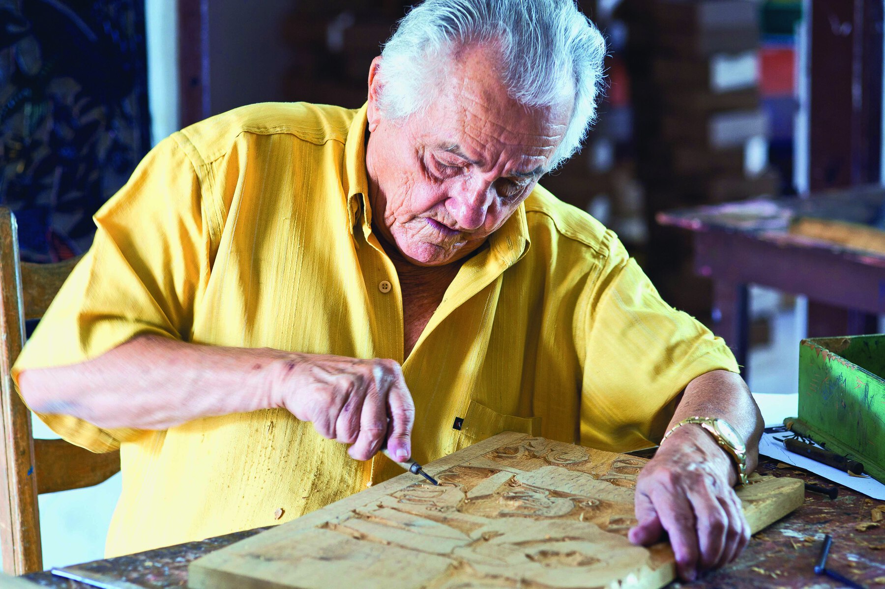 Fotografia. Homem idoso, de cabelo grisalho curto, vestindo camisa amarela. Com a mão direita, ele utiliza um formão para fazer entalhes em uma placa de madeira.