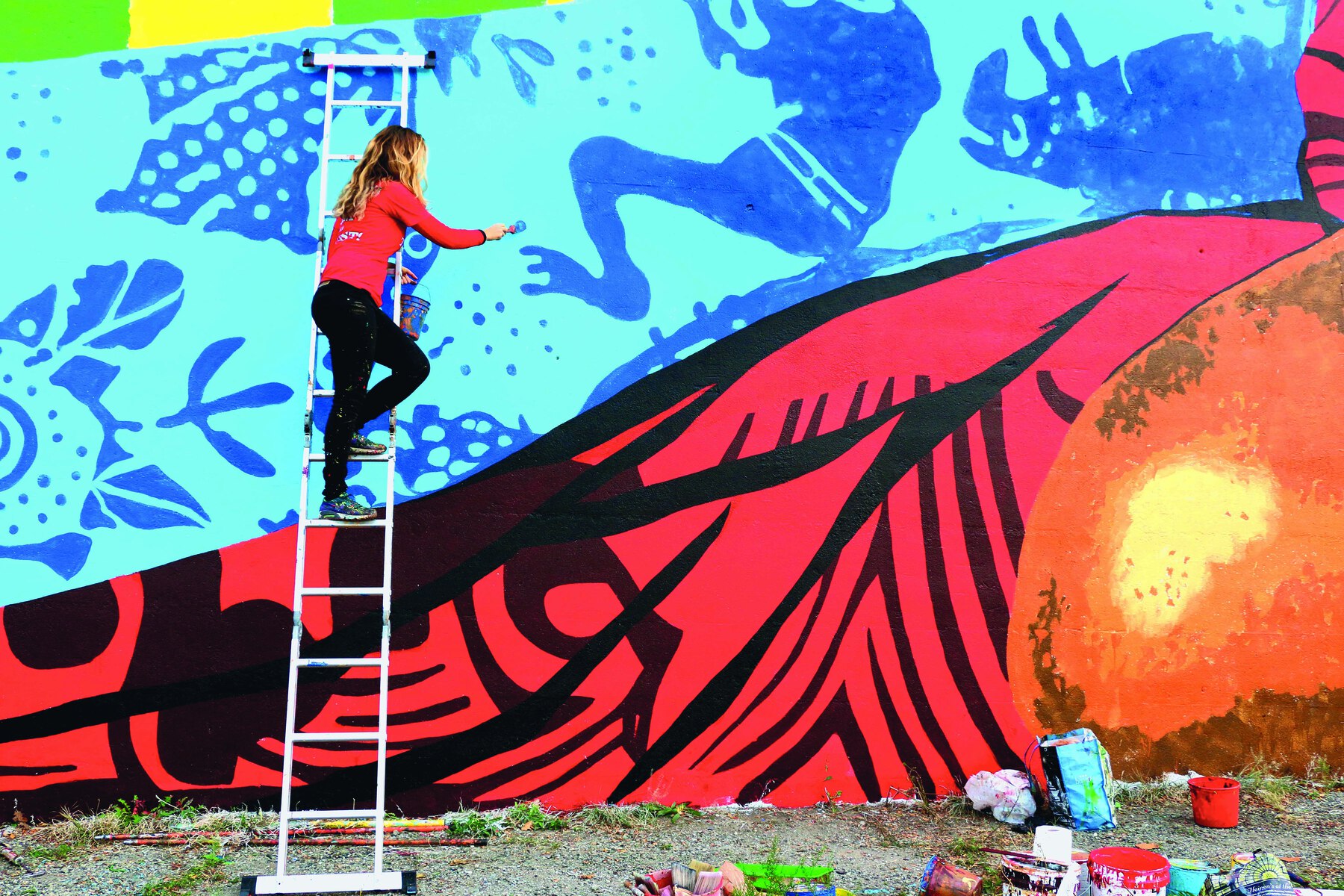 Fotografia. Mulher de cabelo longo loiro, vestindo blusa laranja e calça preta. Está no alto de uma escada pintando um mural em tons de azul com sombras azuis-escuras de animais.