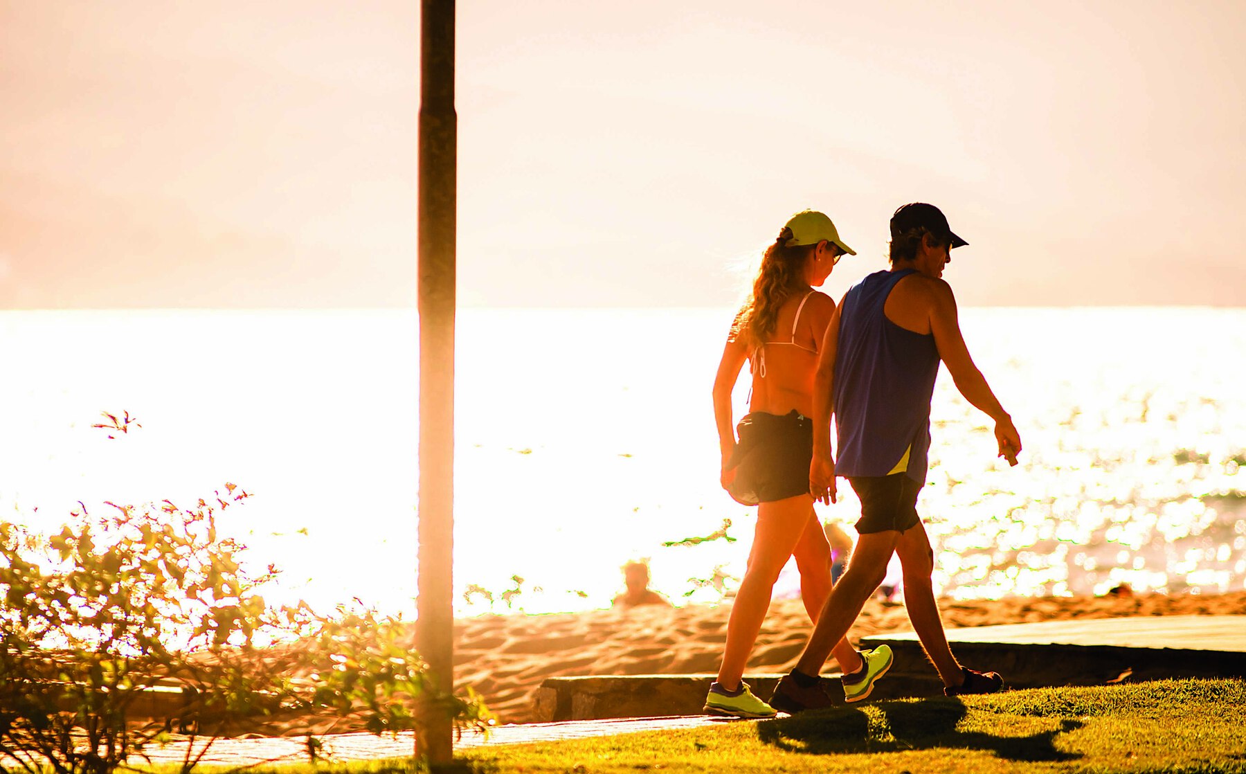 Fotografia. Casal de adultos jovens  fazendo caminhada na calçada da orla de uma praia. Estão quase de costas. A mulher tem cabelo castanho, longo e usa boné, camiseta de alças e short. Ao seu lado, o homem tem cabelo preto, curto e boné. Veste camiseta regata de cor escura e bermuda preta. Ambos estão calçados com tênis. Ao fundo, depois de uma estreita faixa de areia está o mar que, refletindo a luz forte do sol, ilumina toda a paisagem com tons de branco, amarelo muito claro, laranjas, marrons.