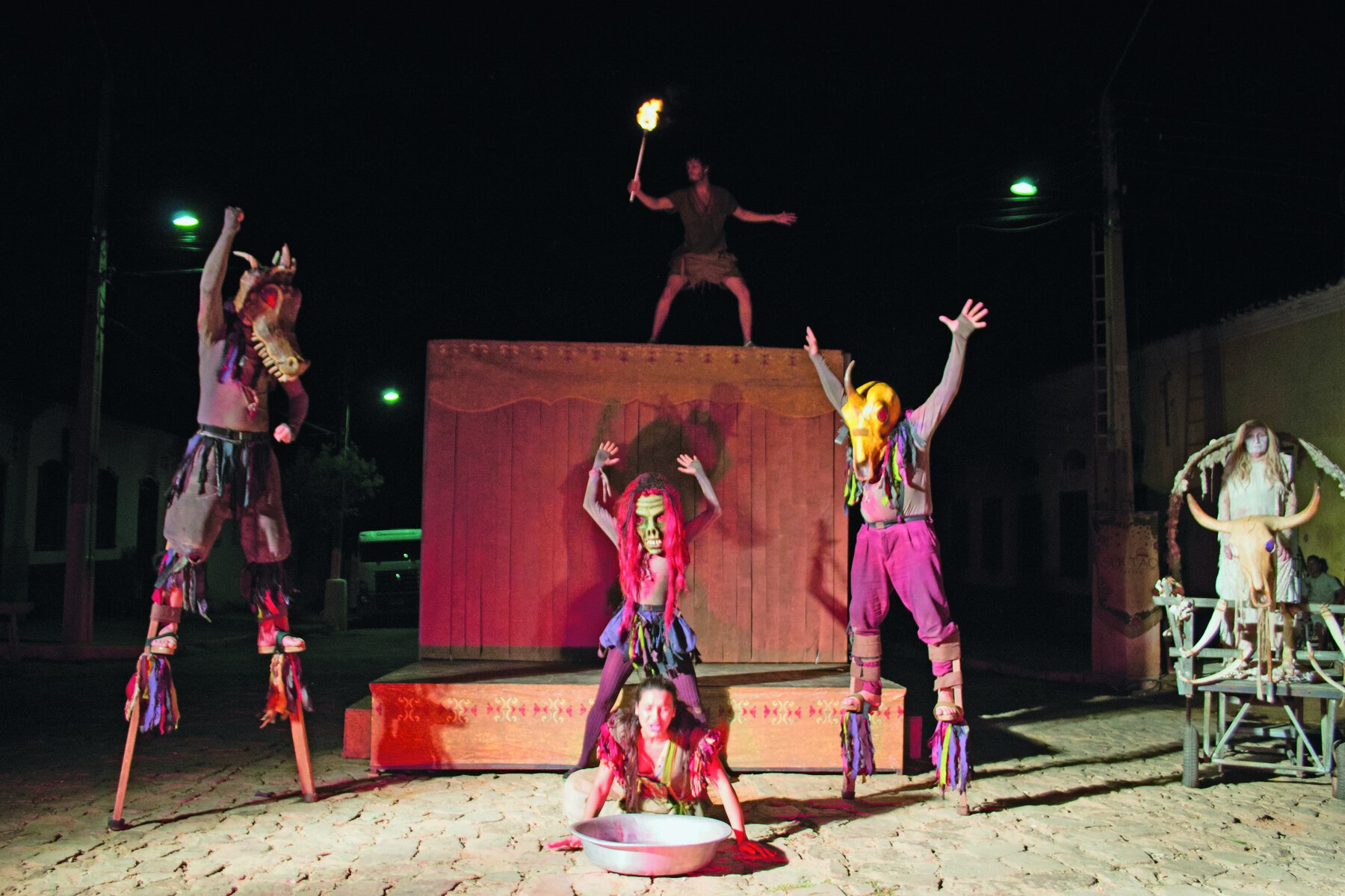 Fotografia. Palco com dois personagens em pernas de pau, um à esquerda da cena, outro à direita. Atrás deles, um suporte de madeira retangular em formato de palco. Um homem segurando uma tocha acesa está no alto ao fundo do suporte.. À frente, ao nível do chão e no centro da imagem, personagem de peruca rosa, com as mãos erguidas. Na frente dela, agachada, uma mulher de cabelo preso segura uma bacia que está apoiada no chão. À direita da cena, mulher de cabelo longo loiro, está sobre a estrutura de uma carroça puxada por uma cabeça de boi