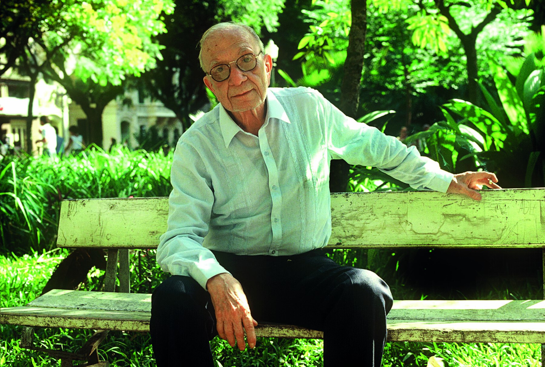 Fotografia. Retrato de homem idoso ao ar livre. Ele é calvo, usa óculos de armação escura, com aros arredondados e finos. Veste camisa verde-clara de mangas compridas. Está sentado em um banco, com a mão esquerda apoiada no encosto de pedra e o tronco ligeiramente inclinado para a frente. Ao fundo aparecem folhagens e árvores.