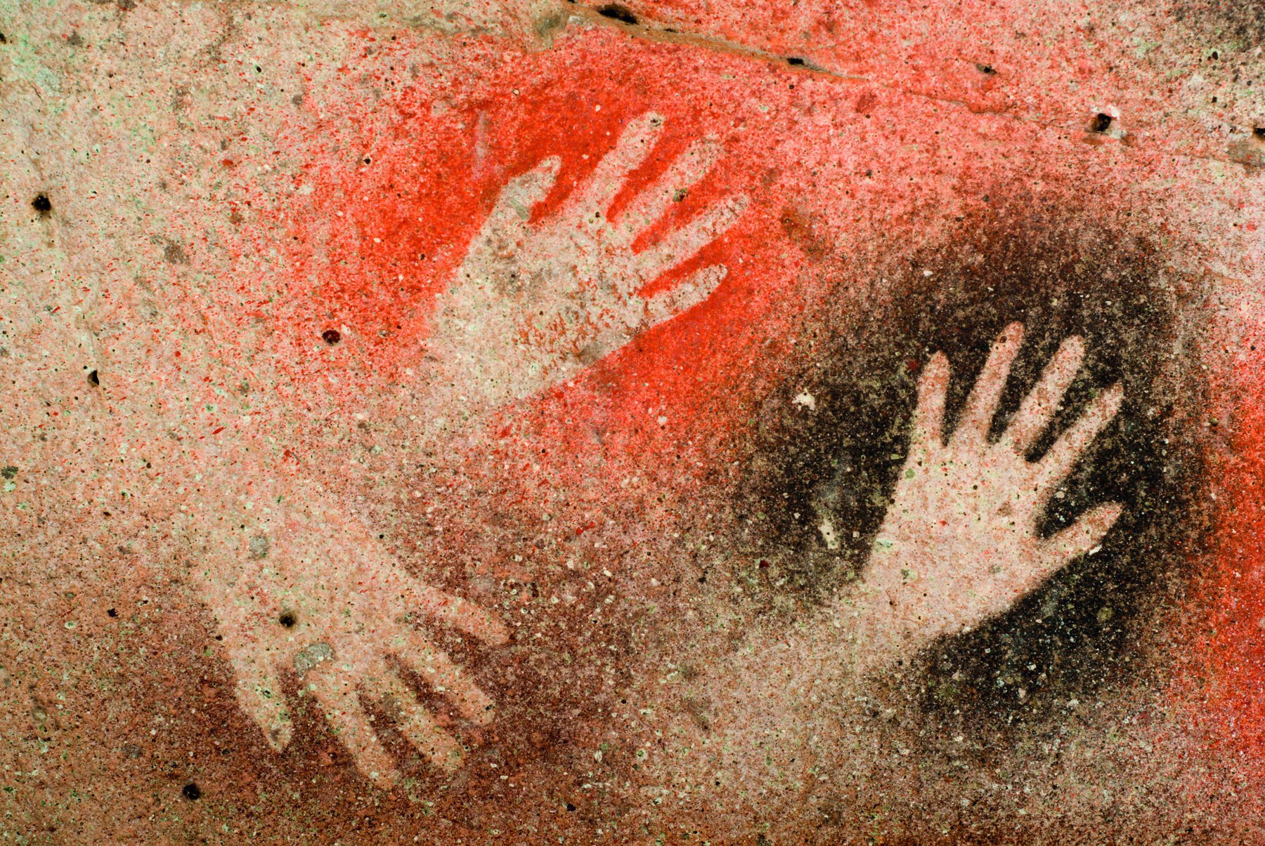 Fotografia. Pintura rupestre mostrando mãos humanas contornadas em tintas de tons de preto, laranja e marrom.