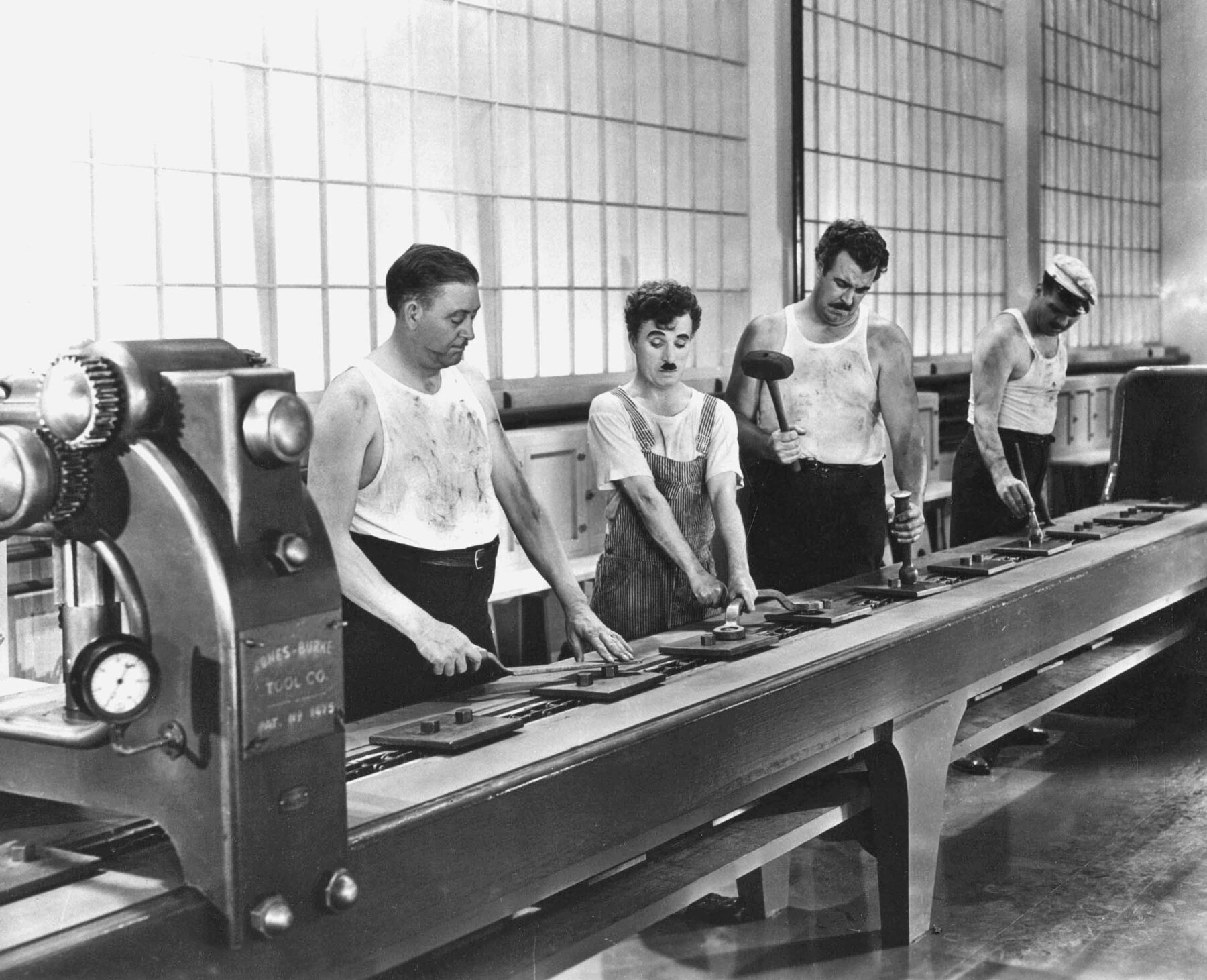 Fotograma em preto e branco. Quatro homens realizam diferentes tarefas na esteira de uma linha de produção de uma fábrica antiga. Três deles vestem camiseta regata branca e calça preta. Da esquerda para a direita, o segundo deles veste camiseta e macacão listrado. O quarto homem usa boné de aba. Todos têm cabelo preto, curto e bigode. À esquerda, em primeiro plano, há a máquina que aciona a linha de produção. Ao fundo, grandes janelas envidraçadas.