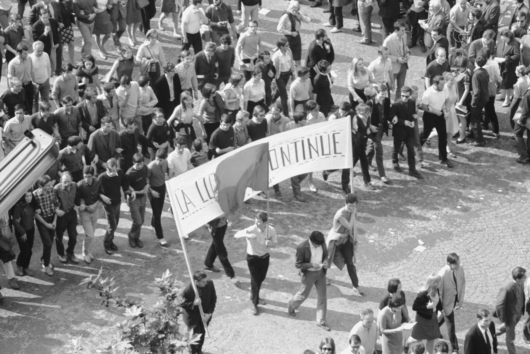 Fotografia em preto e branco. Vista geral de pessoas em uma passeata de protesto. À frente da passeata, dois homens segurando uma faixa grande com palavras de ordem.