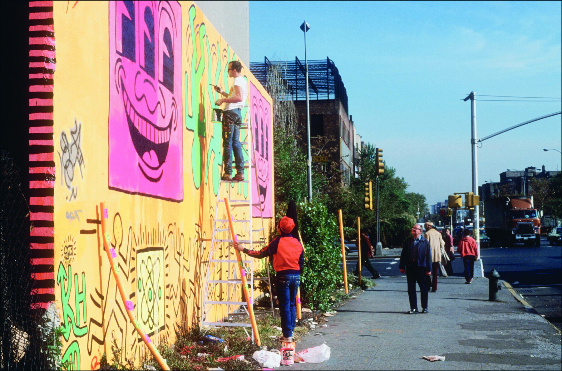 Fotografia. Vista de homem de cabelo curto castanho, vestindo camiseta branca e calça azul. Está no alto de uma escada pintando um mural em tons de verde, amarelo e rosa. No chão, outra pessoa de gorro e blusa cor de laranja segura um cabo de madeira e observa o trabalho.