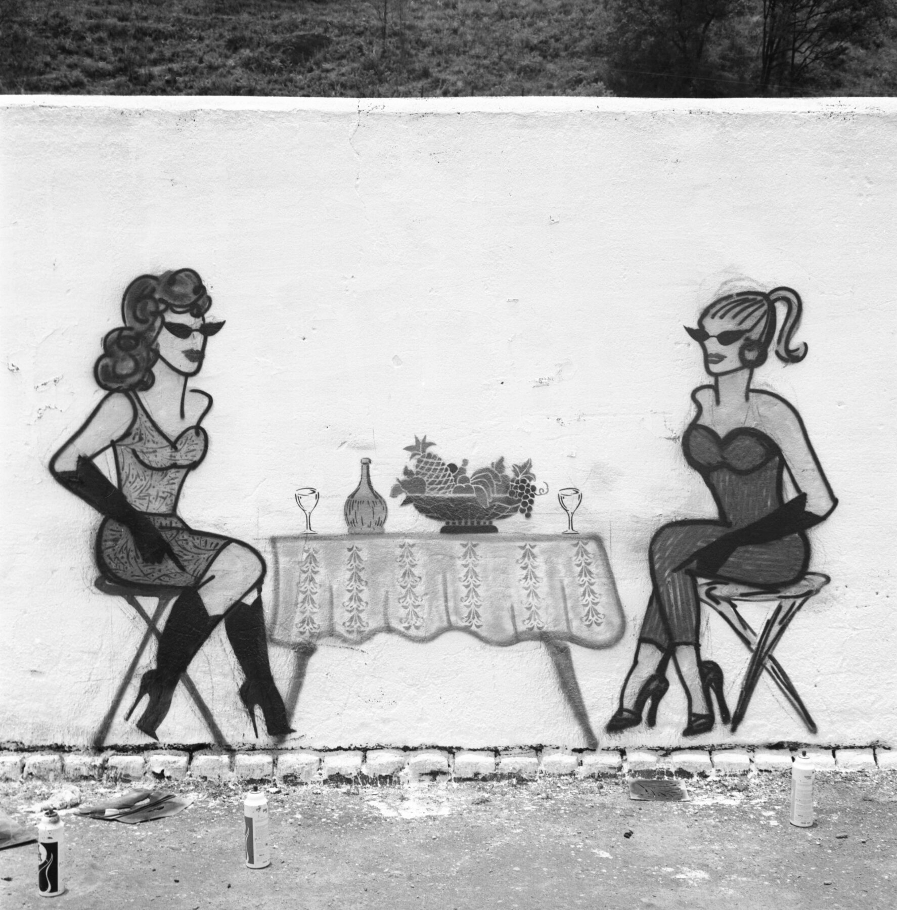 Fotografia em preto e branco. Grafite que mostra duas mulheres de óculos escuros sentadas em torno de uma mesa com frutas em uma fruteira, uma garrafa e duas taças. A mulher da esquerda tem cabelos escuros, está de vestido curto e botas pretas de salto alto. A mulher da direita tem cabelos claros, está de macacão decotado e usa sapatos de salto alto.