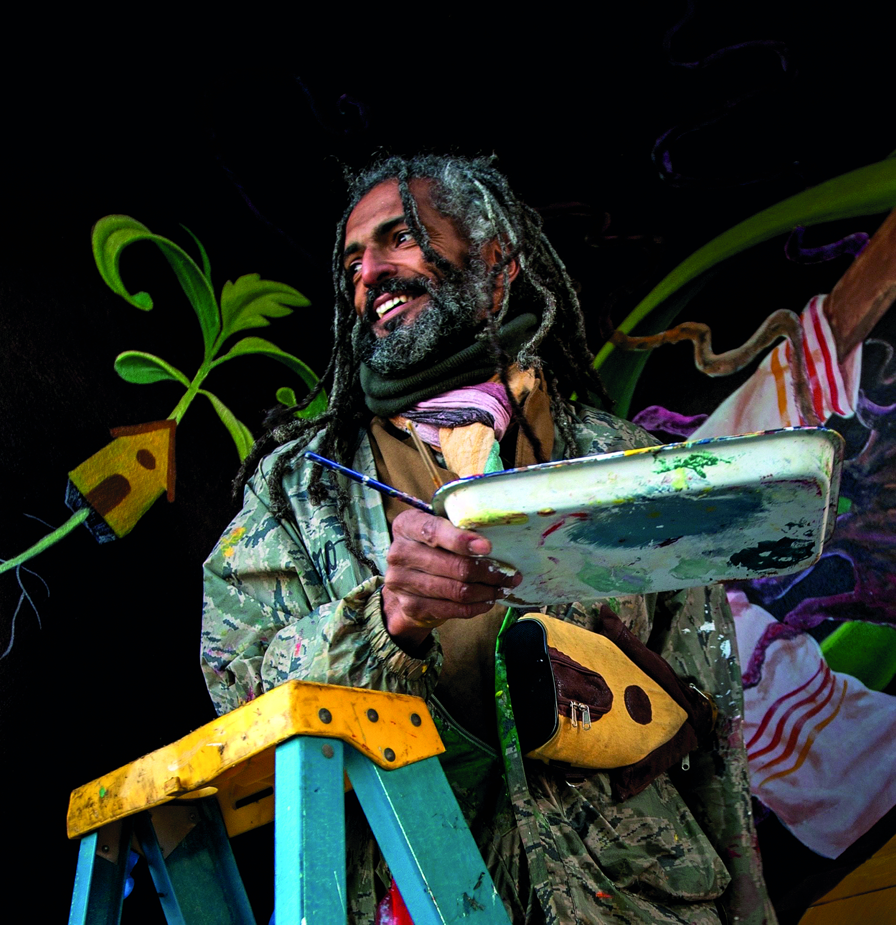 Fotografia. Homem de cabelo, barba e bigode grisalhos vestindo camiseta marrom e jaqueta cinza. Esta sorrindo e segura um pincel e uma bandeja de tinta.