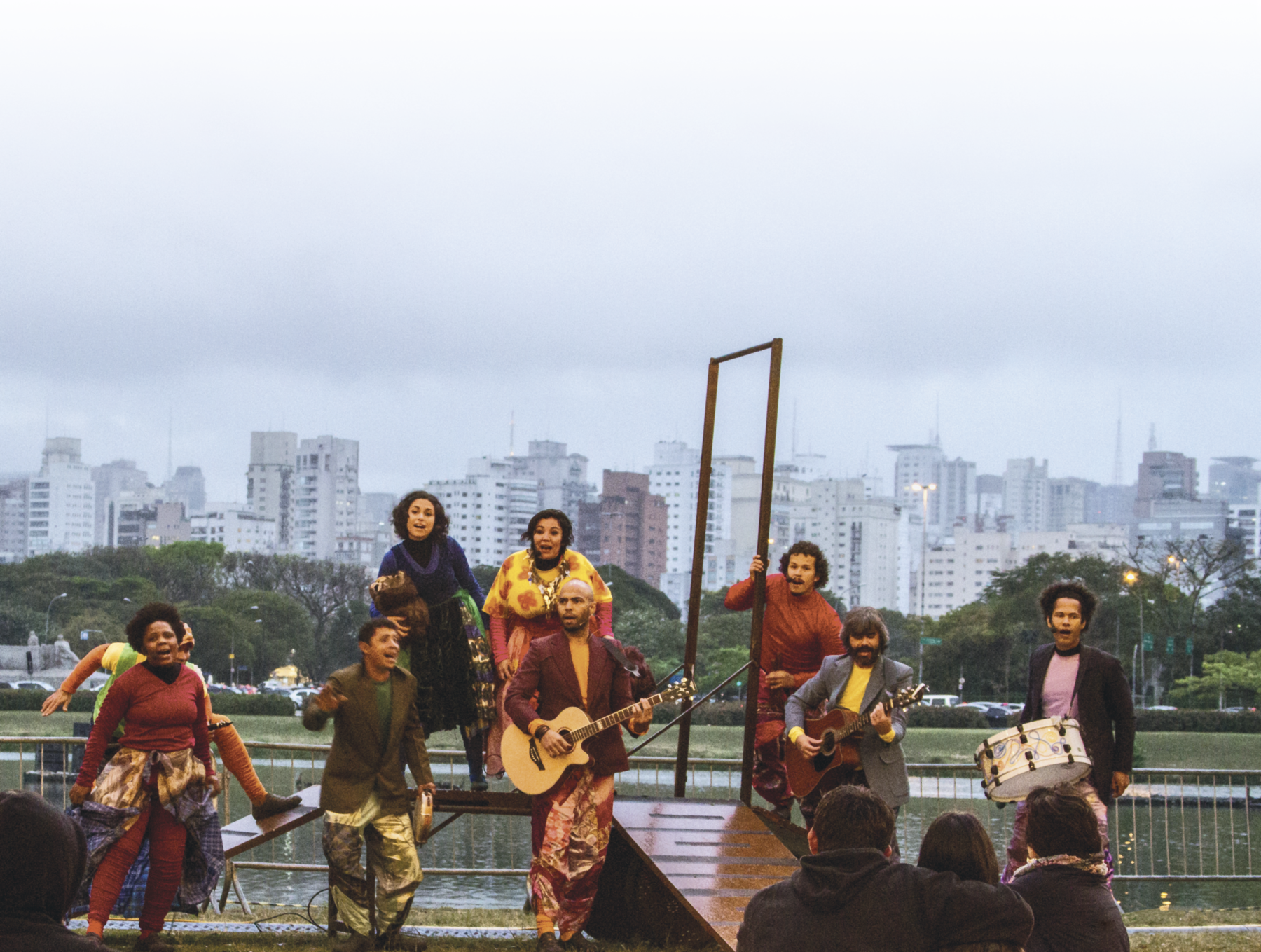 Fotografia. Palco preparado em um ambiente aberto, na frente de um grande lago, com homens e mulheres em uma apresentação musical. Três mulheres e dois homens cantam, enquanto dois homens tocam violão e um toca um tambor. De frente para eles, um grupo de pessoas que parou para observar a cena.