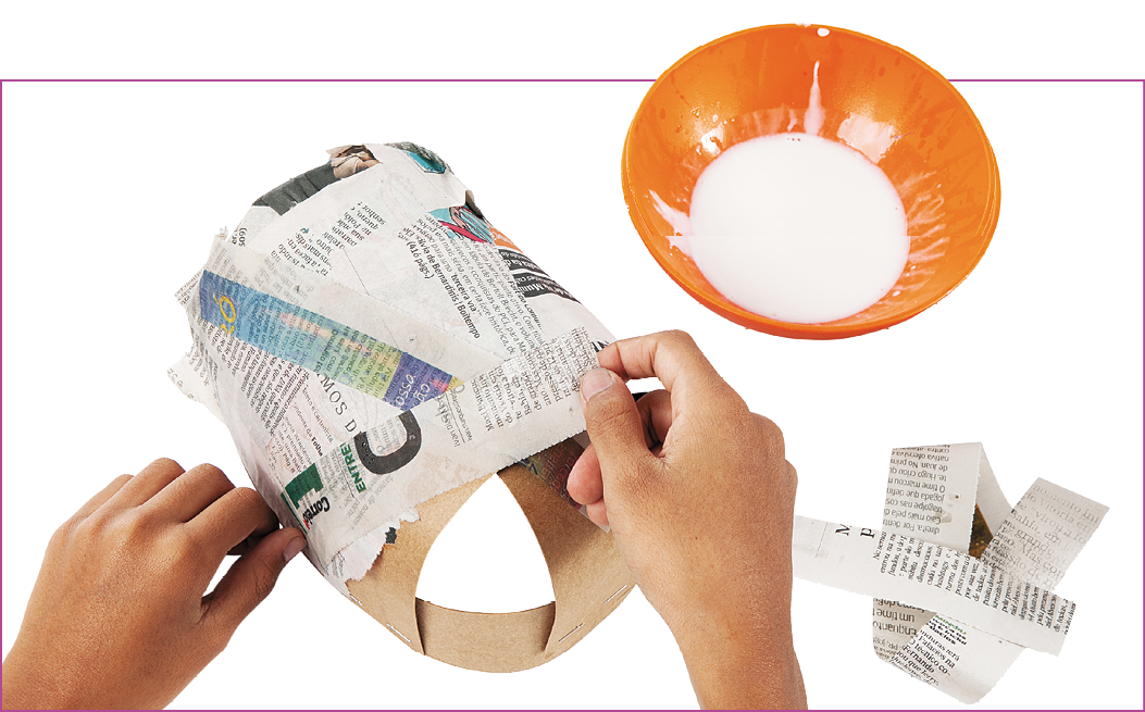 Fotografia. À direita, uma tigela cor de laranja com cola branca . À esquerda,  duas mãos colando tiras de jornal na estrutura de papel-cartão para recobri-la.