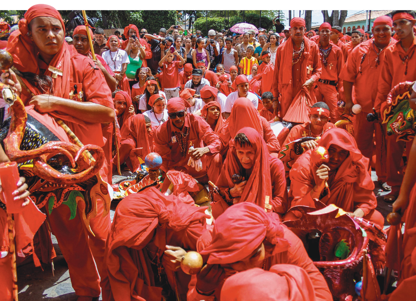 Fotografia. Homens reunidos em um tipo de procissão, vestindo roupas vermelhas e turbantes vermelhos sobre a cabeça. Levam máscaras de demônios nas mãos.