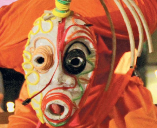 Fotografia. Personagem com máscara de feições distorcidas, com os olhos destacados, um preto e um laranja, e linhas formando pinturas faciais que vão da testa até o redor da boca.