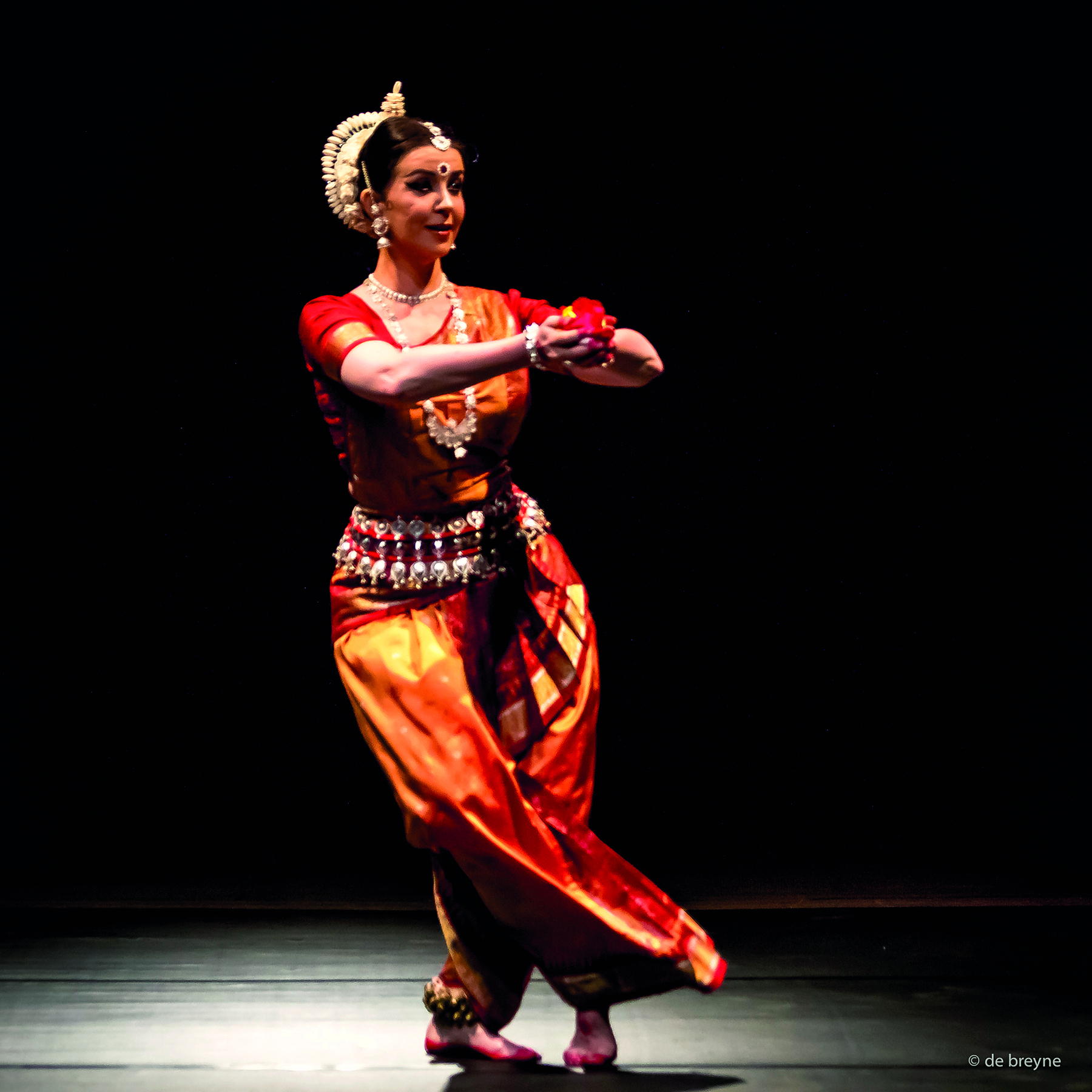 Fotografia. Apresentação de dança indiana. Mulher de cabelo castanho, preso e com um adorno de tom claro na parte posterior da cabeça, realiza passo de dança. Ela está descalça e, com a perna direita cruzada à frente da esquerda, toca o chão com a ponta do pé. O pé esquerdo, mais atrás, está todo apoiado no chão. Os braços estão abertos, cotovelos flexionados, com as duas mãos unidas à frente, na altura do peito, segurando uma flor vermelha. A bailarina veste roupas em estilo indiano: blusa justa de mangas curtas e calça acinturada e com as pernas bufantes, tudo em tons de vermelho e laranja. Ela usa adornos como brincos, colares, pulseiras grandes e cinto de contas.