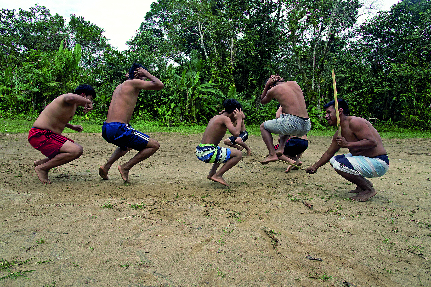 Fotografia. Cinco homens e dois meninos indígenas realizando uma dança em um terreno plano, de areia. Há vegetação variada, verde, ocupando todo o fundo. Os homens usam apenas bermudas e estão descalços, realizando movimentos de agachar e pular, enquanto se movimentam em círculo. As mãos estão apoiada nas laterais da cabeça. Um dos homens está mais à direita, agachado, fora do círculo de dançarinos. Com expressão sorridente, segura uma vara de madeira em cada uma das mãos.