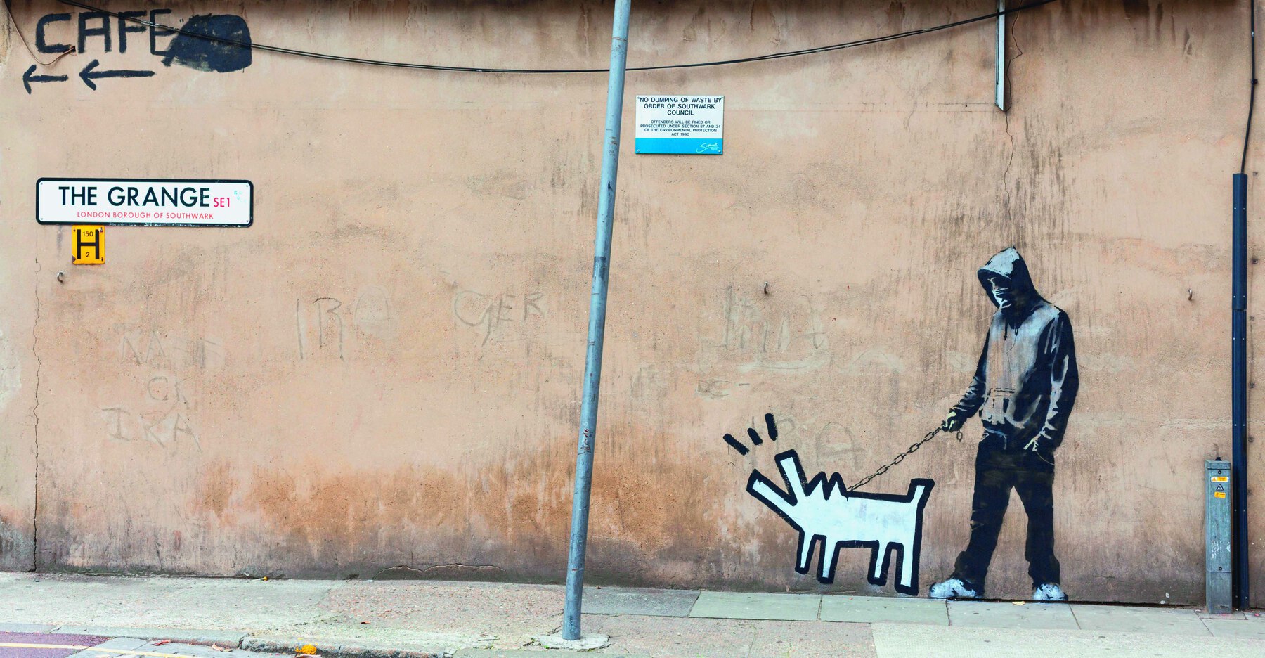 Fotografia. Grafite sobre uma parede: homem com blusa de capuz cinzenta, calça escura e tênis brancos, segura uma corrente que vai até a silhueta de um cachorro, desenhado somente com linhas geométricas.