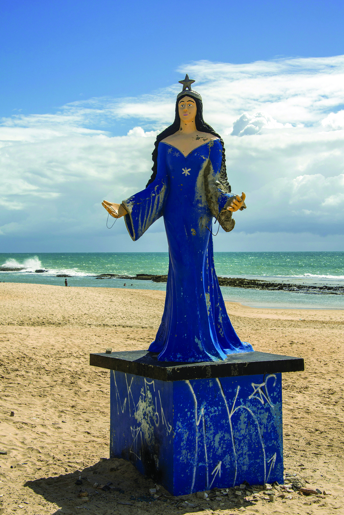 Fotografia. Estátua de figura feminina com cabelos castanhos longos, coroa e vestido azul longo. Está sobre um pedestal, em uma praia, com o mar ao fundo.