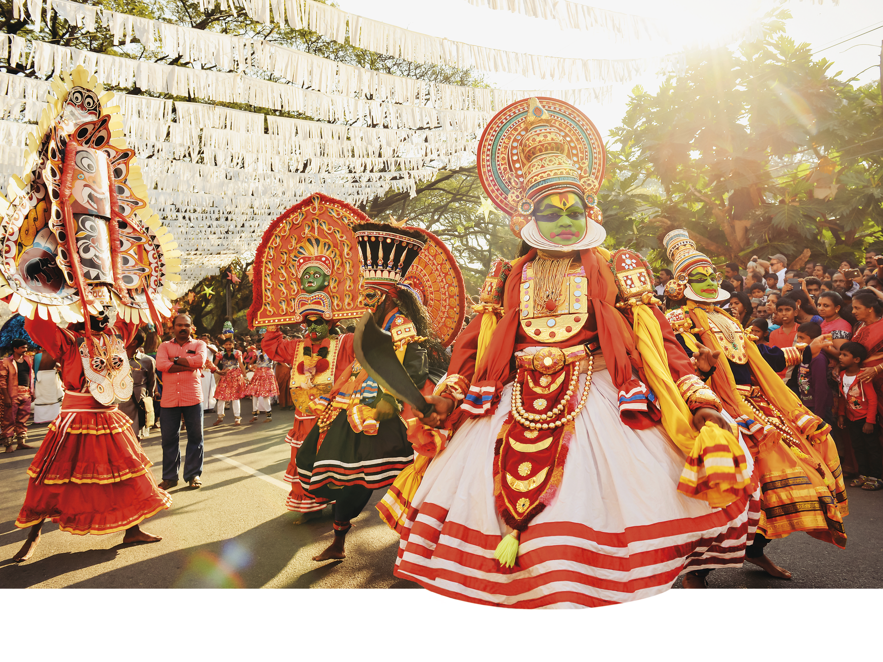 Fotografia. Apresentação de dança indiana ao ar livre. Os intérpretes usam chapéus grandes ornamentados em tons de laranja, vermelho, amarelo e preto e vestem-se nos mesmos tons. As saias são compridas e rodadas, com listras coloridas na barra.  Na pintura facial  predomina o tom verde, com detalhes amarelos. À esquerda, uma figura de vestimenta vermelha, longa, usa na cabeça uma grande máscara de várias faces. No fundo da cena, à esquerda e à direita, uma multidão assiste à apresentação.