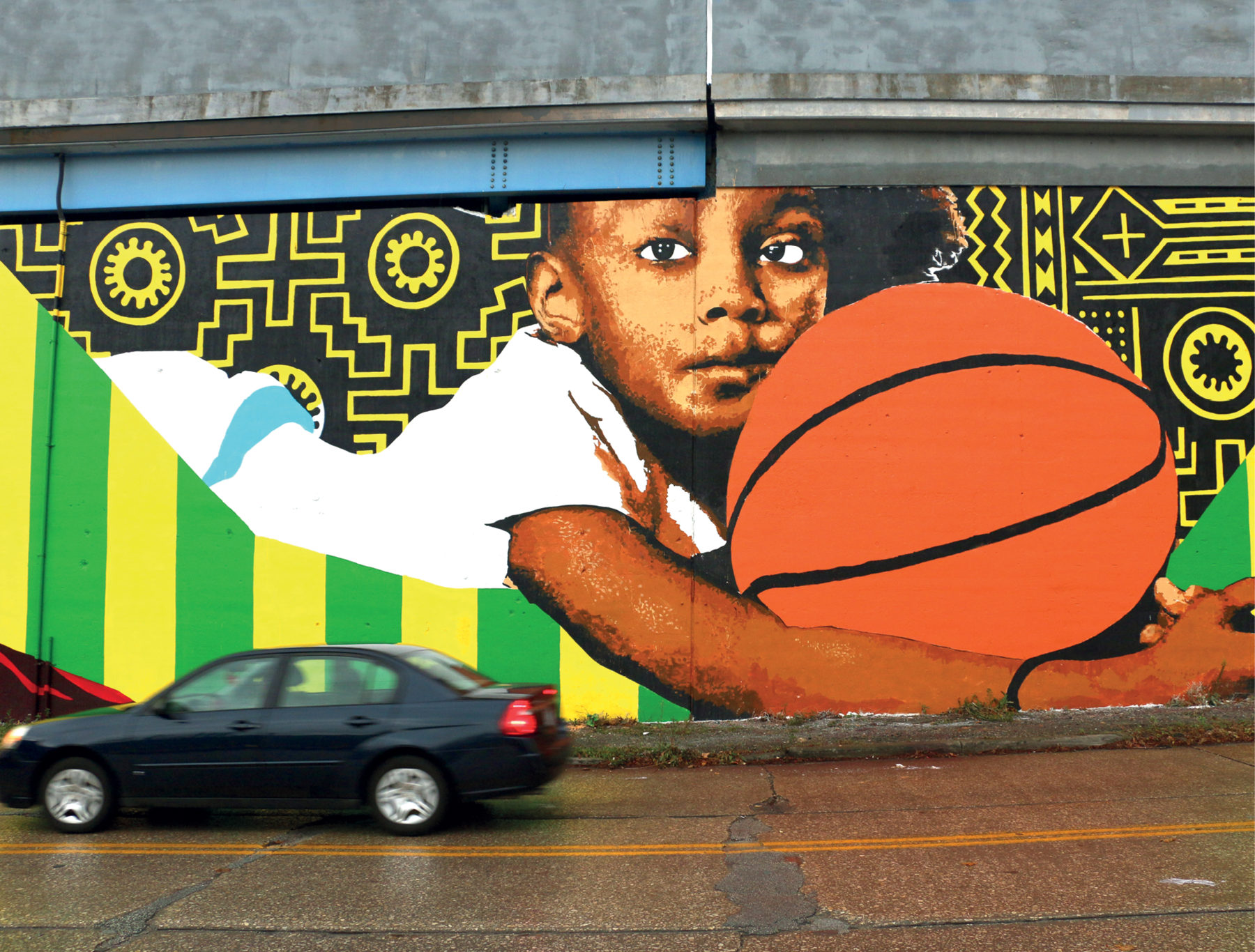 Fotografia. Detalhe de pintura mural, mostrando um menino de cabelo curto preto. Ele veste camiseta branca e está deitado de barriga para baixo, segurando uma bola de basquete. Ao redor do menino há desenhos simétricos em preto e amarelo e listras verdes e amarelas.