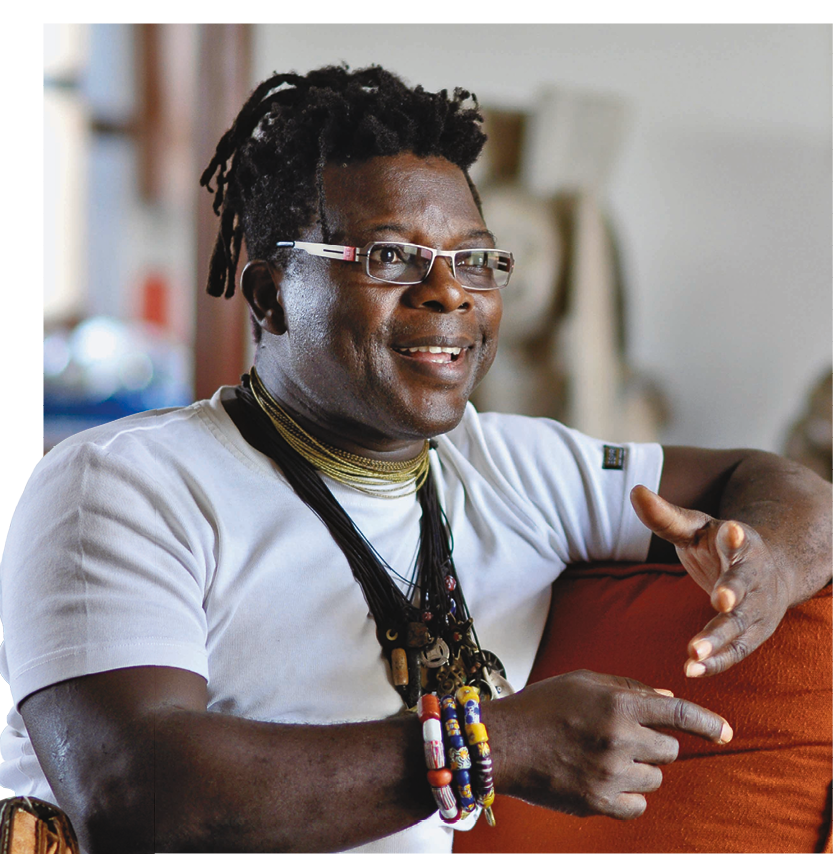 Fotografia. Homem negro de cabelo preto, médio, todo trançado. Usa óculos de armação retangular, veste camiseta branca, colares e pulseiras de contas e sementes coloridas. Tem expressão sorridente.