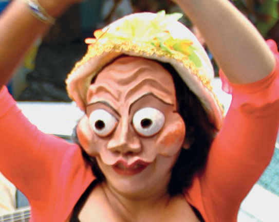 Fotografia. Personagem com uma máscara de olhos arregalados e saltados e bochechas proeminentes. Ela usa um chapéu amarelo adornado de verde-claro. Uma blusa cor de laranja cobre os braços até os cotovelos.