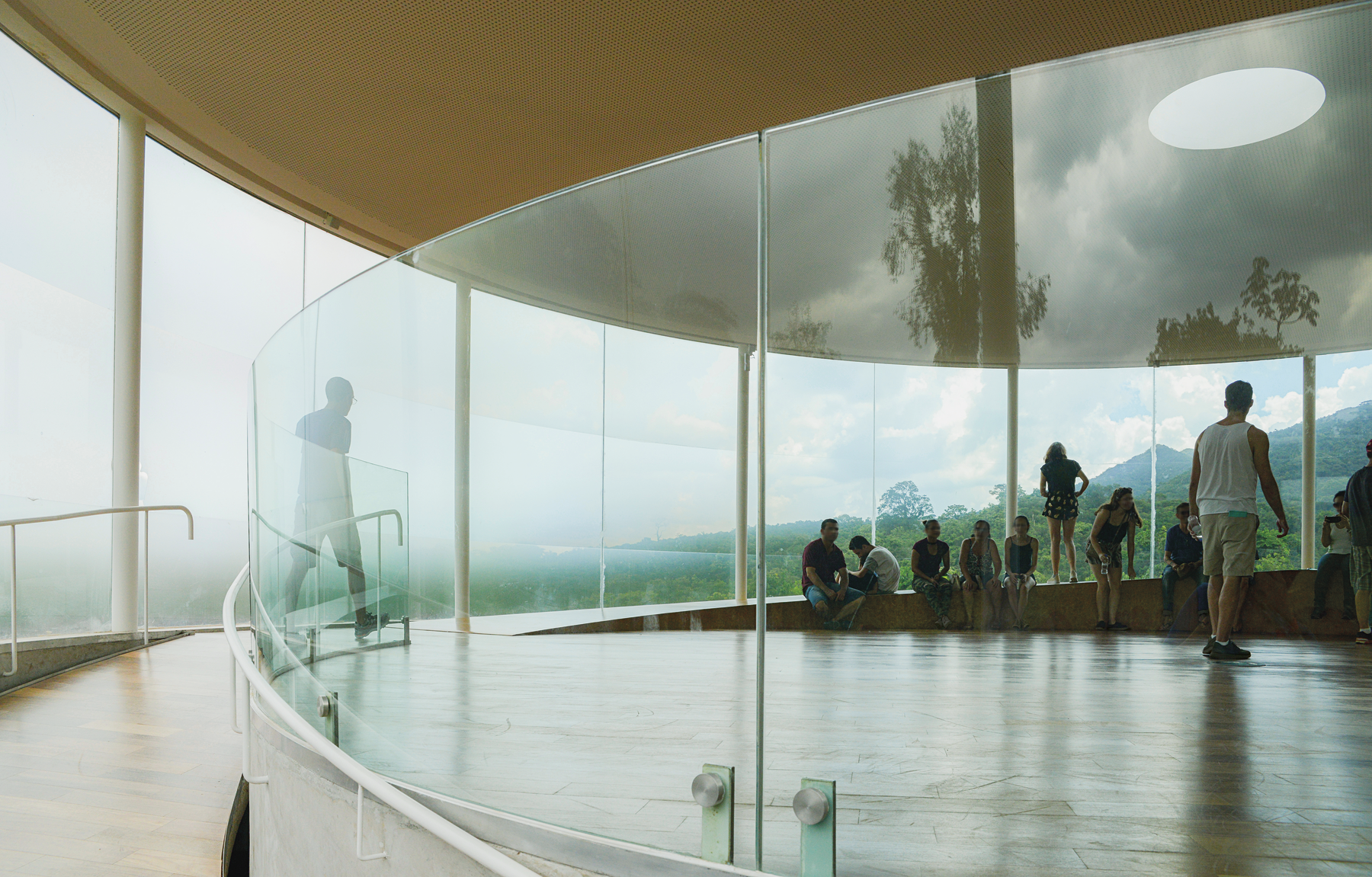 Fotografia. Interior de um pavilhão circular com paredes de vidro e vegetação na área externa. Dentro dele, algumas pessoas estão sentadas, e outras estão em pé.