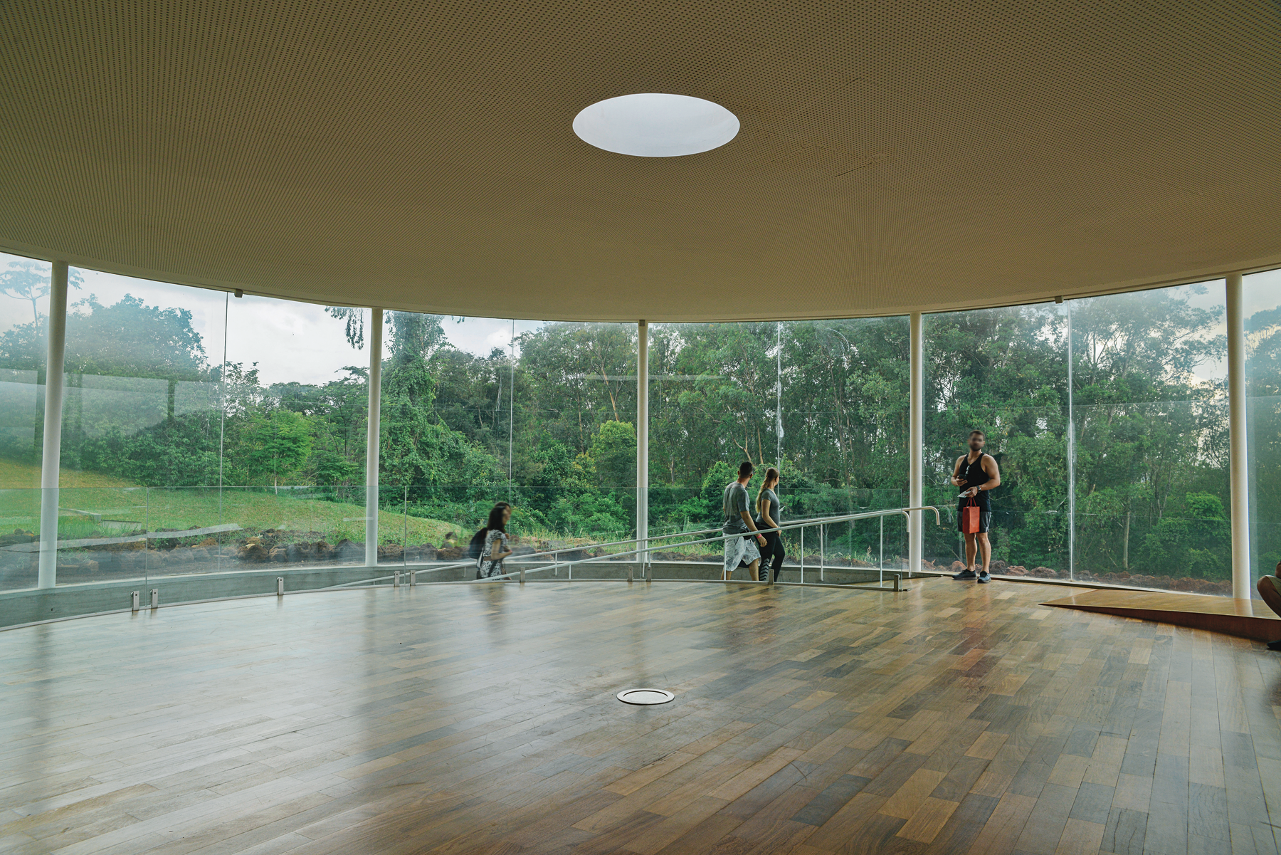 Fotografia. Interior de um pavilhão circular com paredes de vidro e vegetação na área externa. O salão tem piso de madeira, e uma rampa com corrimão. Há pessoas descendo pela rampa.