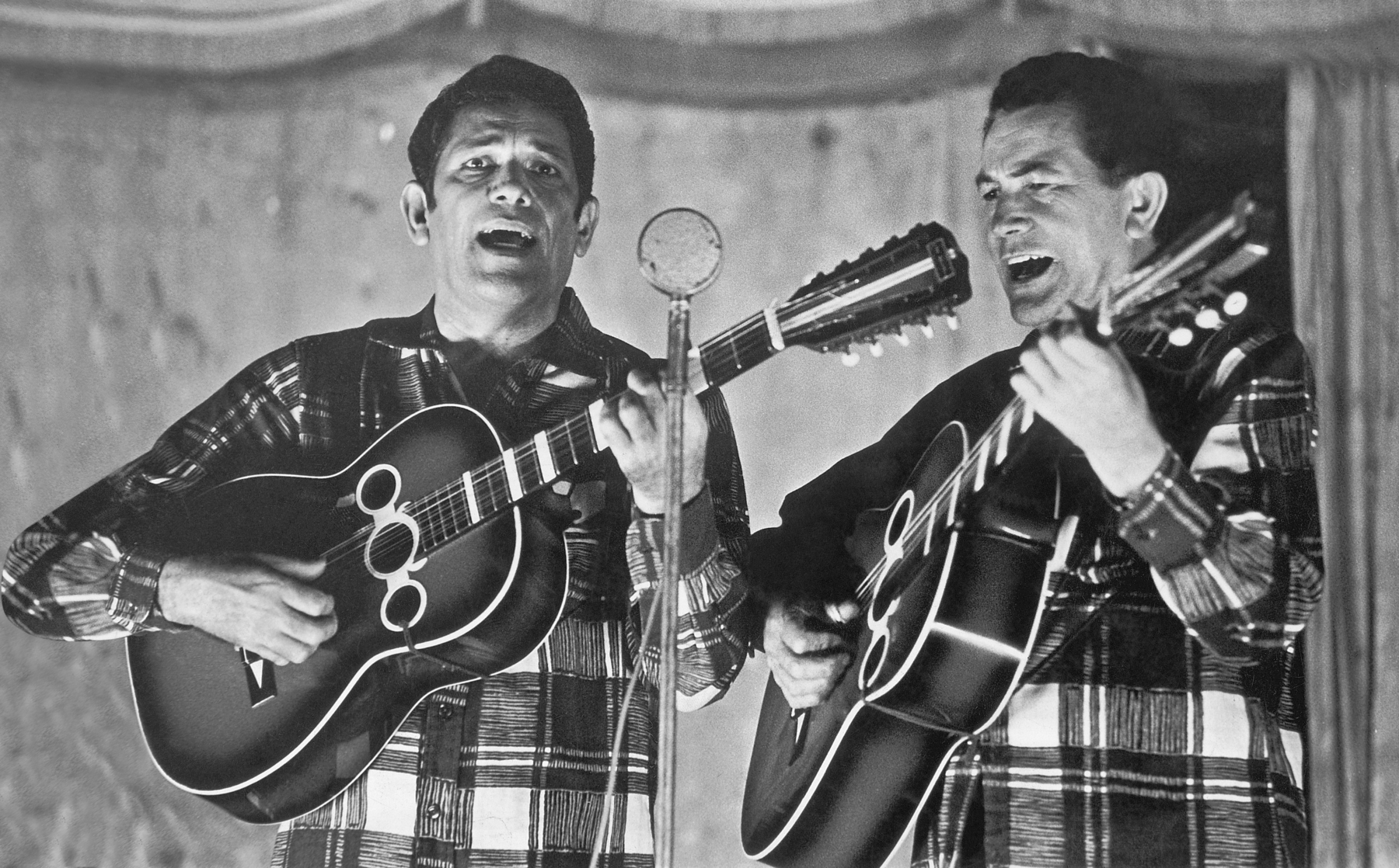 Fotografia em preto e branco. Dois homens brancos e de cabelos curtos, vestindo camisa xadrez e tocando violão, com a boca aberta. Diante eles há um microfone.