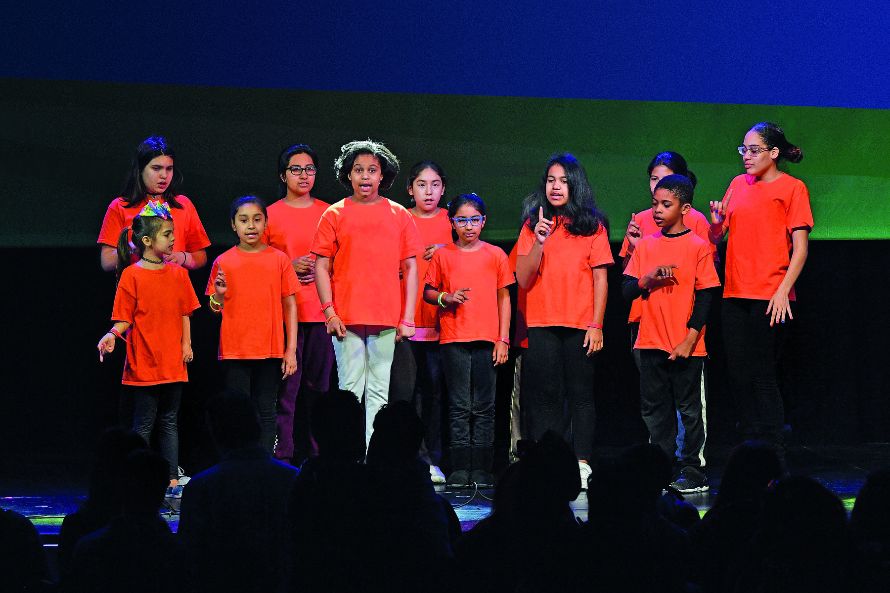 Fotografia. Vários adolescentes sobre um palco vestem camiseta laranja em frente a uma plateia.