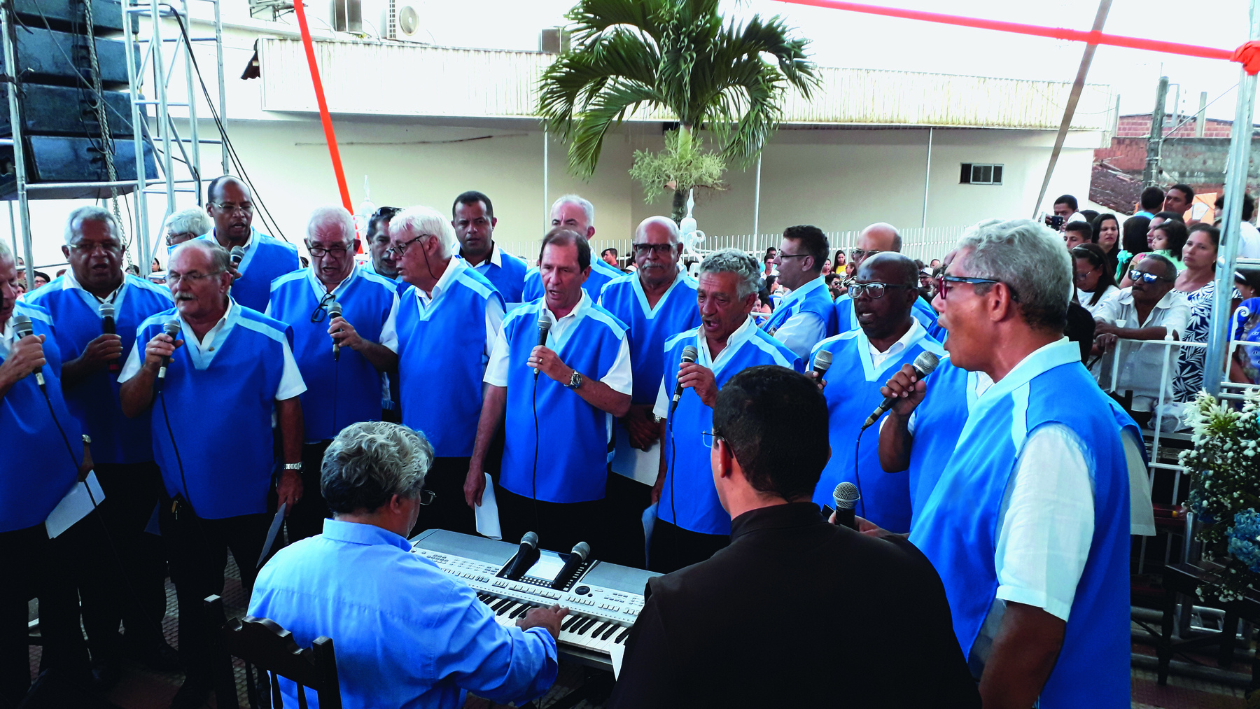 Fotografia. Homens idosos de camisa branca e colete azul. Eles cantam em microfones e estão em pé lado a lado em torno de um homem idoso de cabelo grisalho de camisa azul que está sentado e toca um teclado.