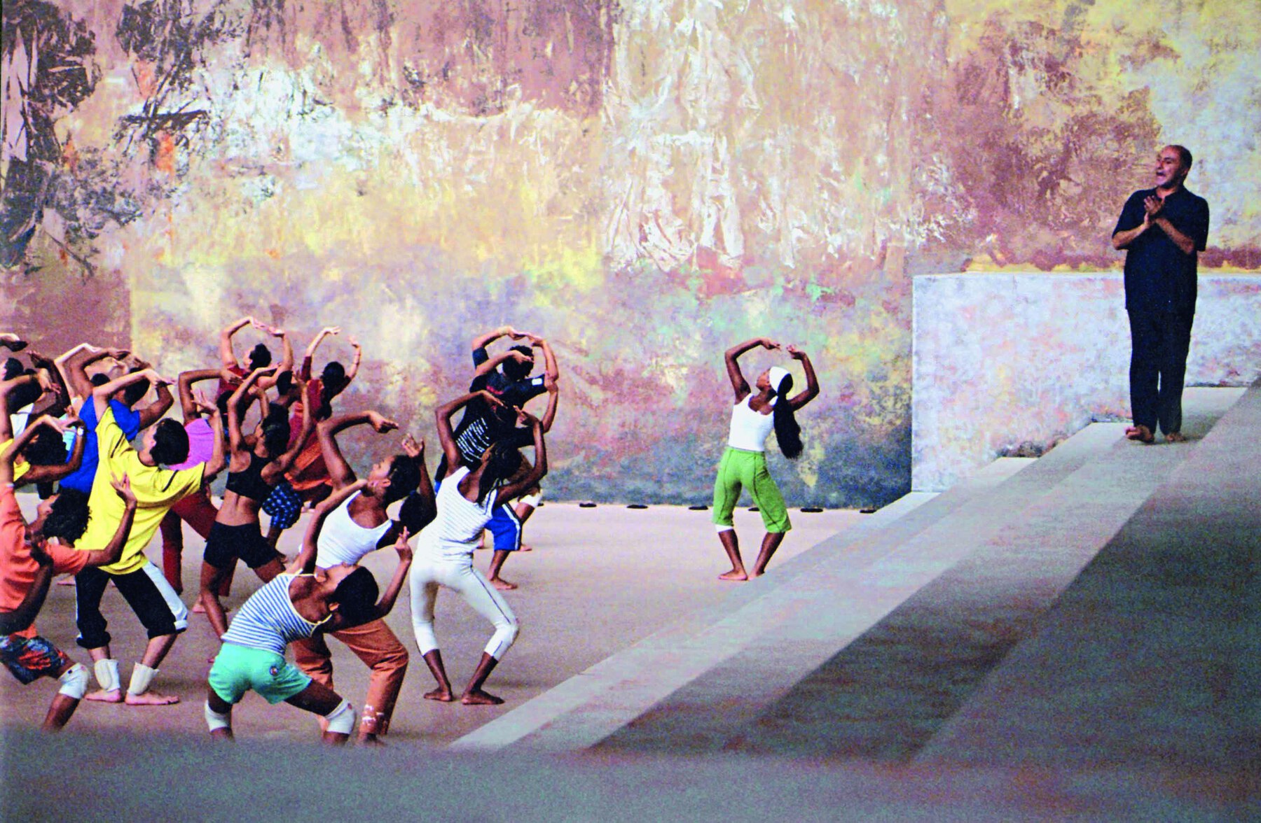 Fotografia. Grupo de dança realizando movimentos com joelhos flexionados e mãos formando arco acima da cabeça. Estão em um salão amplo e fechado, orientados por um homem vestido de preto que está à direita, sobre degraus altos cinza.