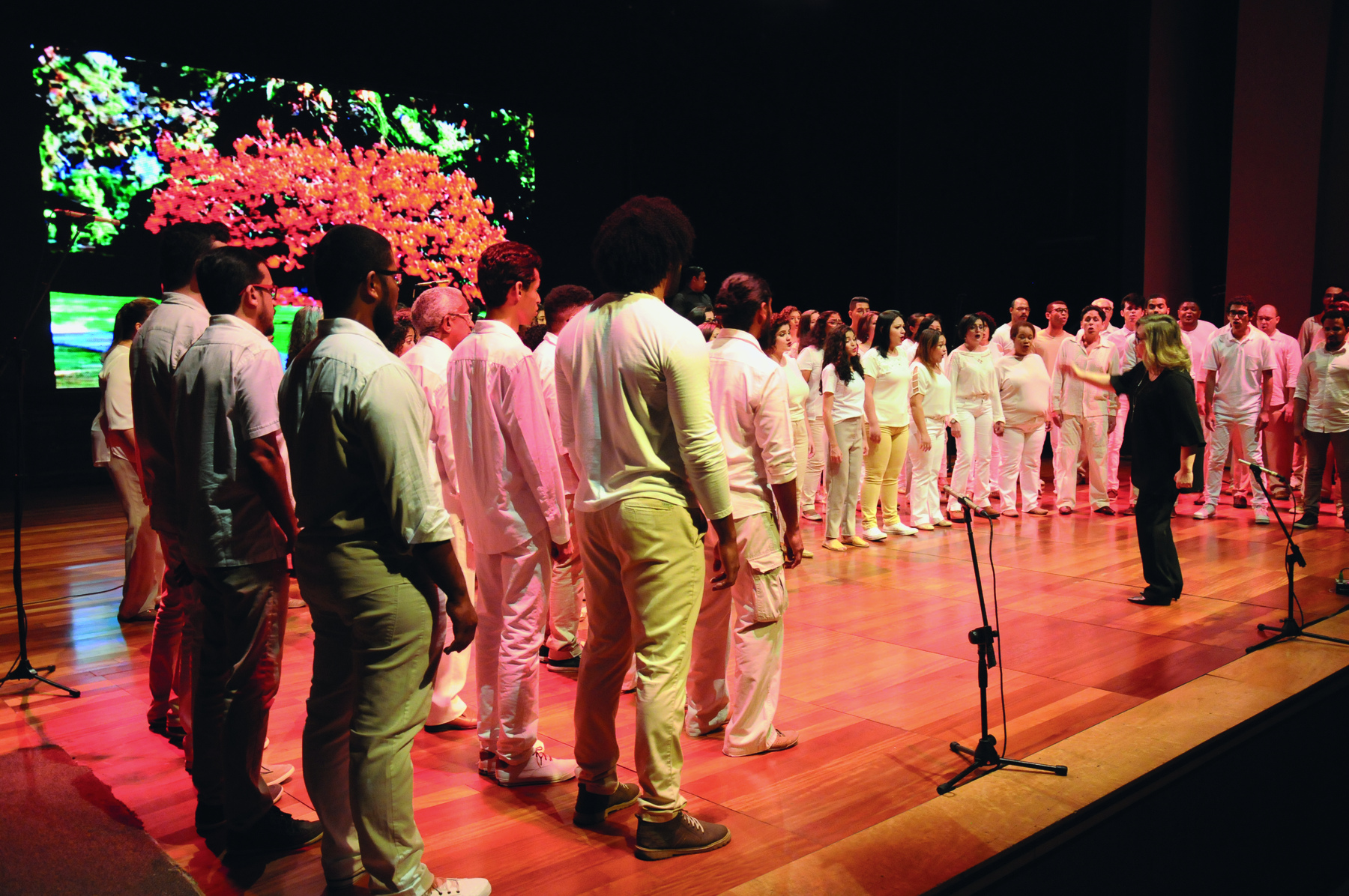 Fotografia. Sobre um palco de madeira clara, homens e mulheres em pé, vestindo calças e blusas brancas, formando um semicírculo. No centro, de frente para as pessoas,  uma mulher de cabelo loiro vestindo camisa e calça preta rege o coral. No meio do palco, um microfone em um pedestal.