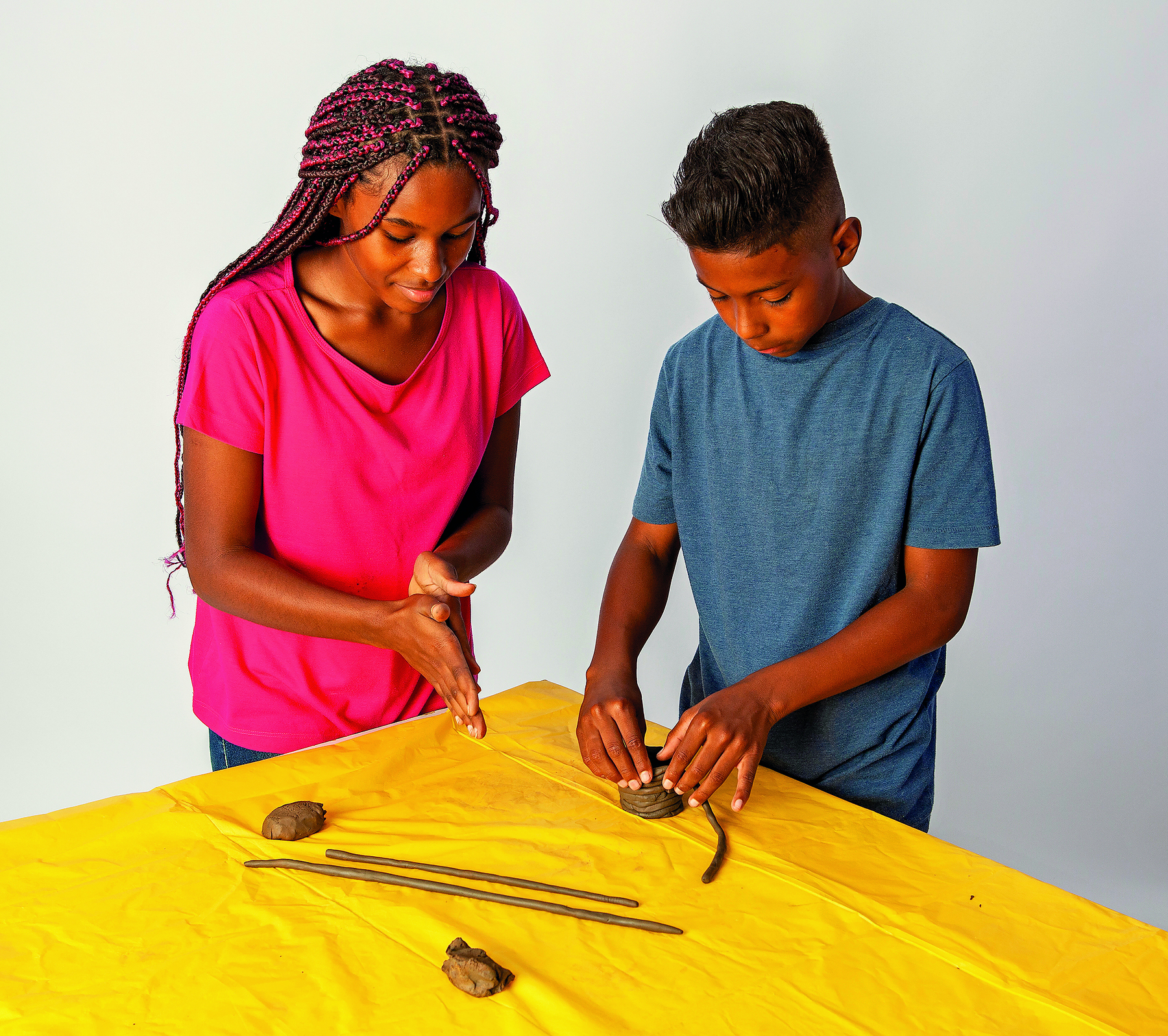 Fotografia. Menina adolescente  com tranças longas em tons de castanho e vermelho, vestindo camiseta rosa. Ao lado dela, menino de cabelo curto preto, vestindo camiseta azul. Eles modelam argila sobre uma mesa coberta por uma toalha amarela.