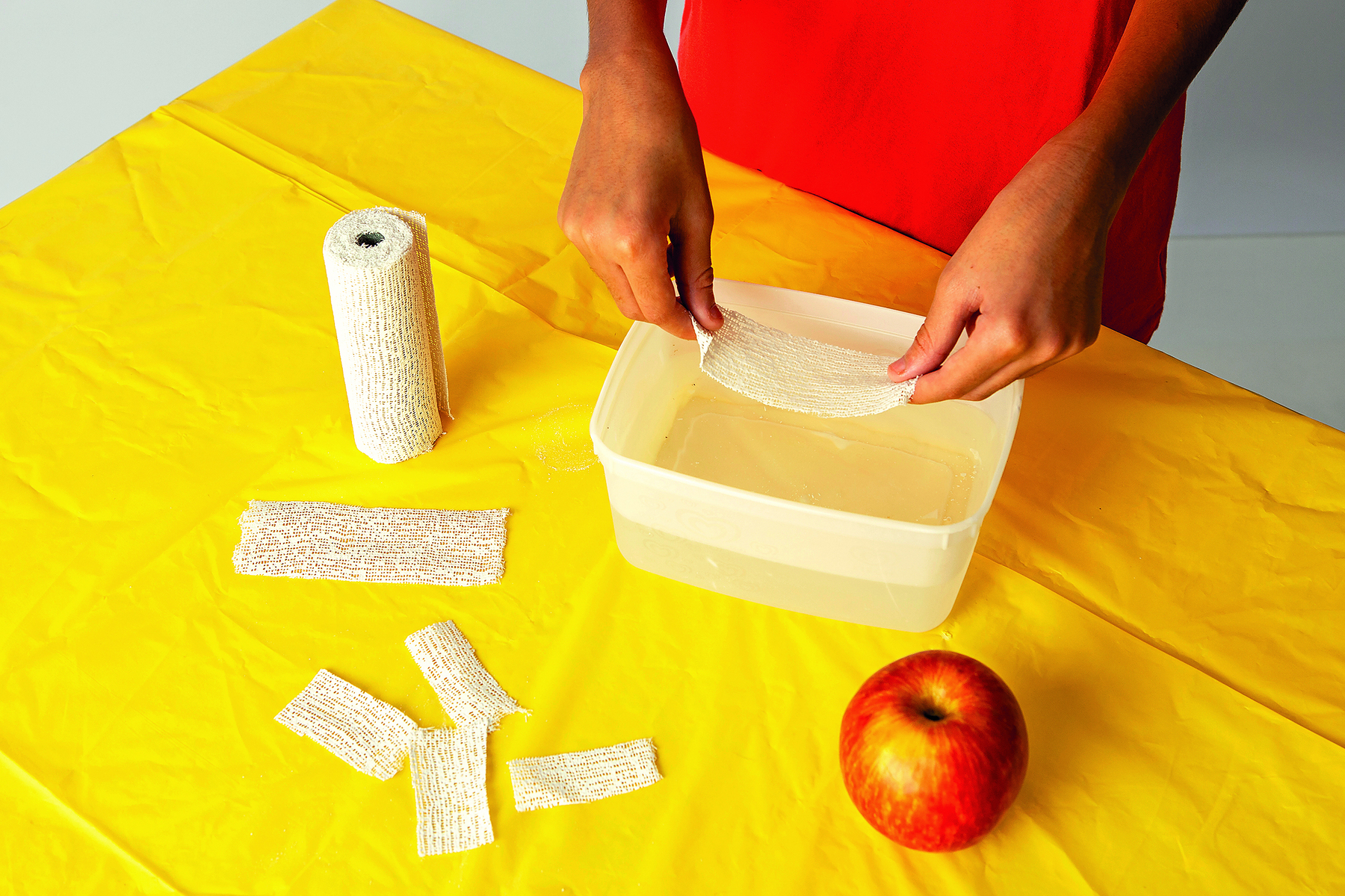 Fotografia. Detalhe de mãos segurando uma atadura acima de um pote com água sobre uma mesa coberta por uma toalha amarela. Ao lado, há um rolo de ataduras, pedaços de ataduras e uma maçã.