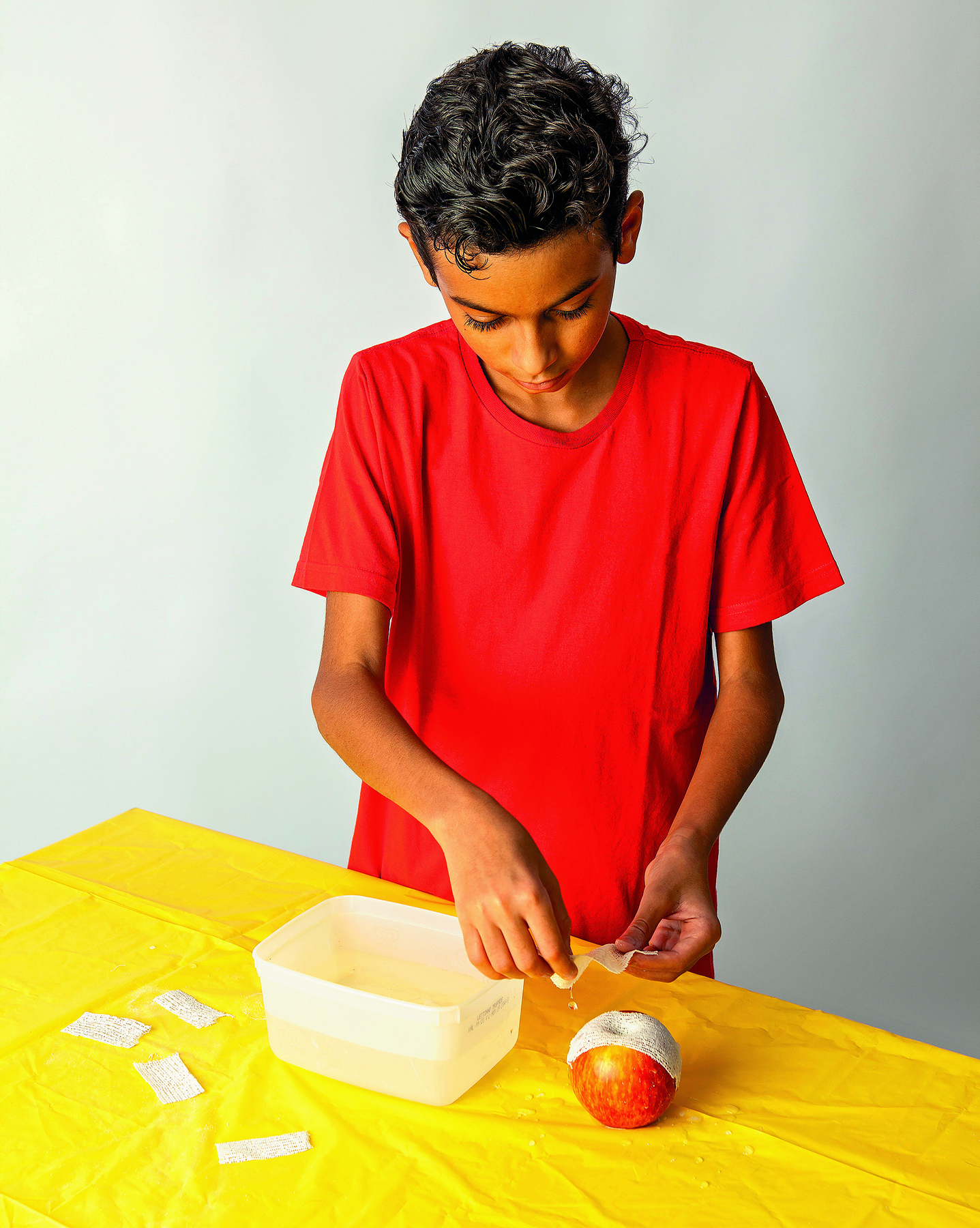 Fotografia. Menino adolescente de cabelo preto, vestindo camiseta vermelha. Com as duas mãos, ele aplica ataduras molhadas em uma  maçã sobre uma mesa coberta por uma toalha amarela, onde há um pote com água e algumas ataduras espalhadas.