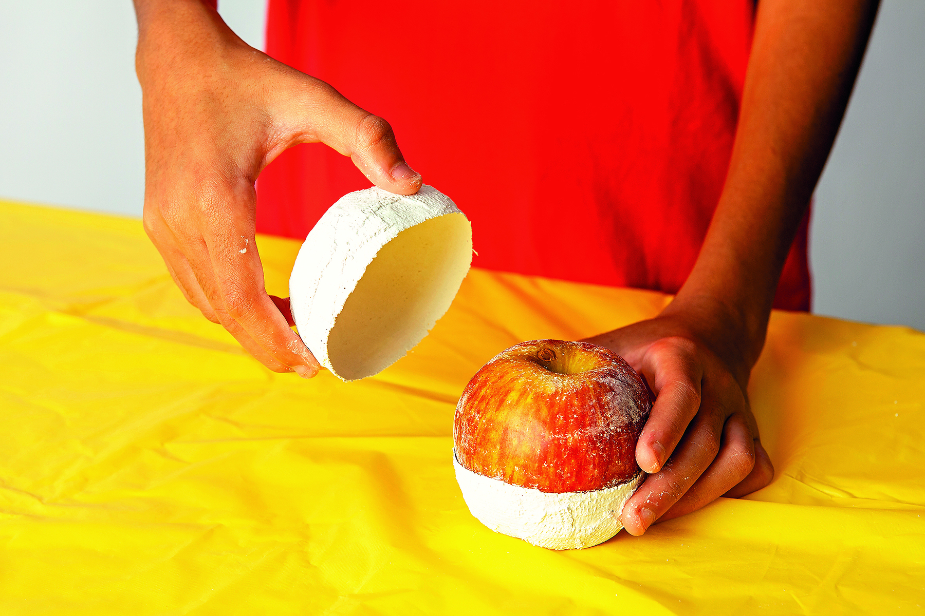 Fotografia. Detalhe das mãos de uma pessoa  removendo a metade superior de um molde de gesso, com a forma de uma maçã, sobre uma mesa coberta por uma toalha amarela.