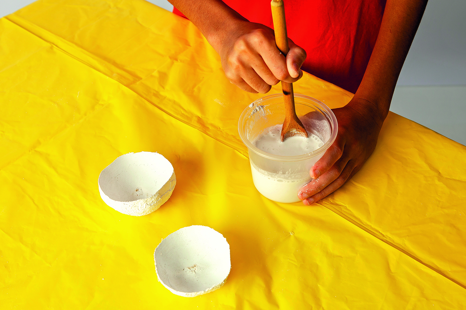 Fotografia. Detalhe de duas maõs mexendo um mistura de gesso em um pote transparente com uma colher de madeira. O pote está apoiado sobre uma mesa coberta por uma toalha amarela, onde há duas metades de um molde de gesso em formato de maçã.
