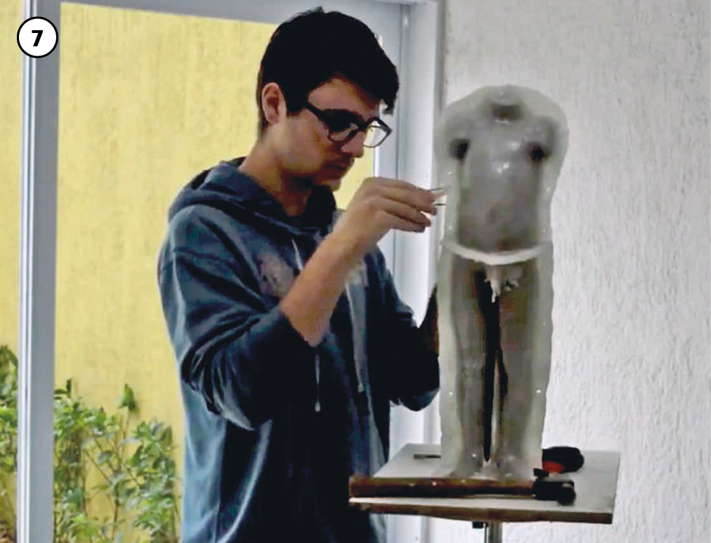 7: O artista, vestido com um blusão azul, retoca a escultura do corpo, que está sem os braços e a cabeça, apoiada em um suporte, sobre uma pequena bancada.