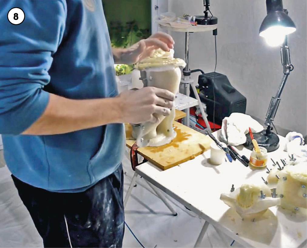 Fotografia. Imagem 8 de uma sequência fotográfica que retrata o processo de produção da escultura de um menino.
Mãos do artista, que veste blusão azul e calça preta com manchas de tinta, modelam uma peça da escultura em um suporte apoiado em uma pequena bancada à frente dele, sobre a qual há uma luminária acesa e diversos instrumentos utilizados pelo escultor.