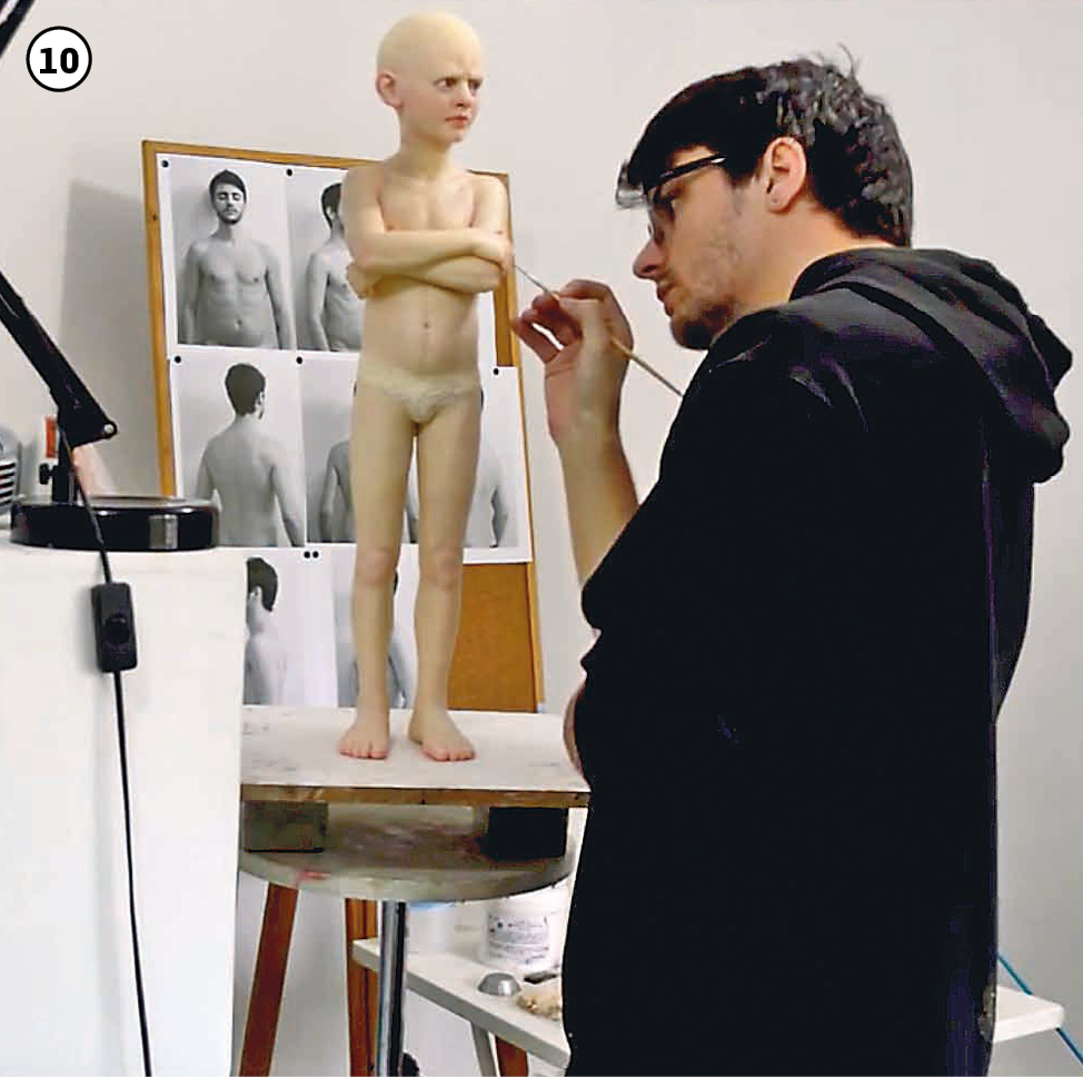 10: O artista, vestido com blusão preto, retoca, com um pincel fino, a escultura de um menino de braços cruzados sobre o peito, que está sobre uma pequena bancada à frente do artista. Ao fundo, há um cavalete com fotografias em preto e branco, em diversos ângulos, de um homem de torso nu, com os braços estendidos ao longo do corpo.