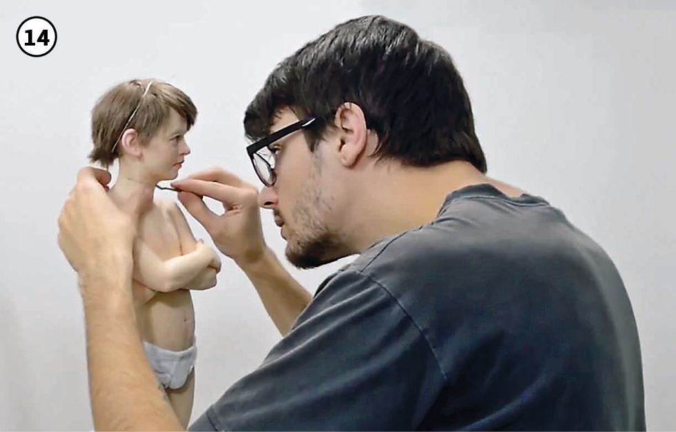 14: O artista, vestido com camiseta cinza, retoca com um pincel fino a conexão da cabeça e do corpo da escultura de um menino, que está com os braços cruzados sobre o peito e veste apenas uma cueca branca.