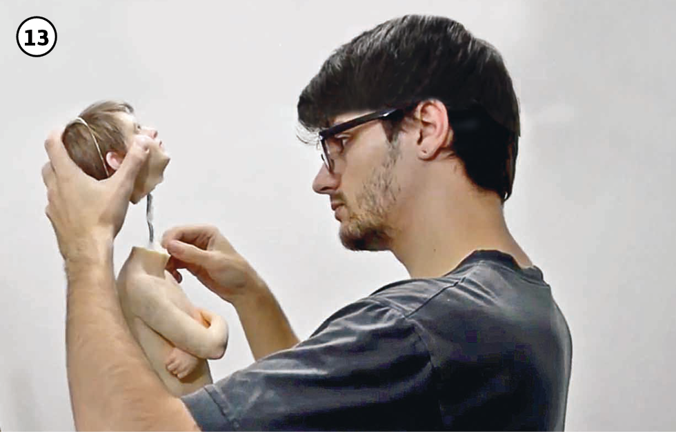 13: O artista, vestido com camiseta cinza, monta as peças da escultura do corpo de um menino de braços cruzados sobre o peito, posicionando a cabeça da figura, já com todos os fios de cabelo nela aplicados.