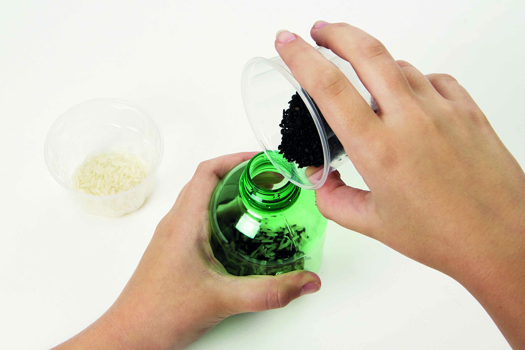 Fotografia. Destaque de mãos despejando sementes pretas dentro de uma garrafa PET pequena.