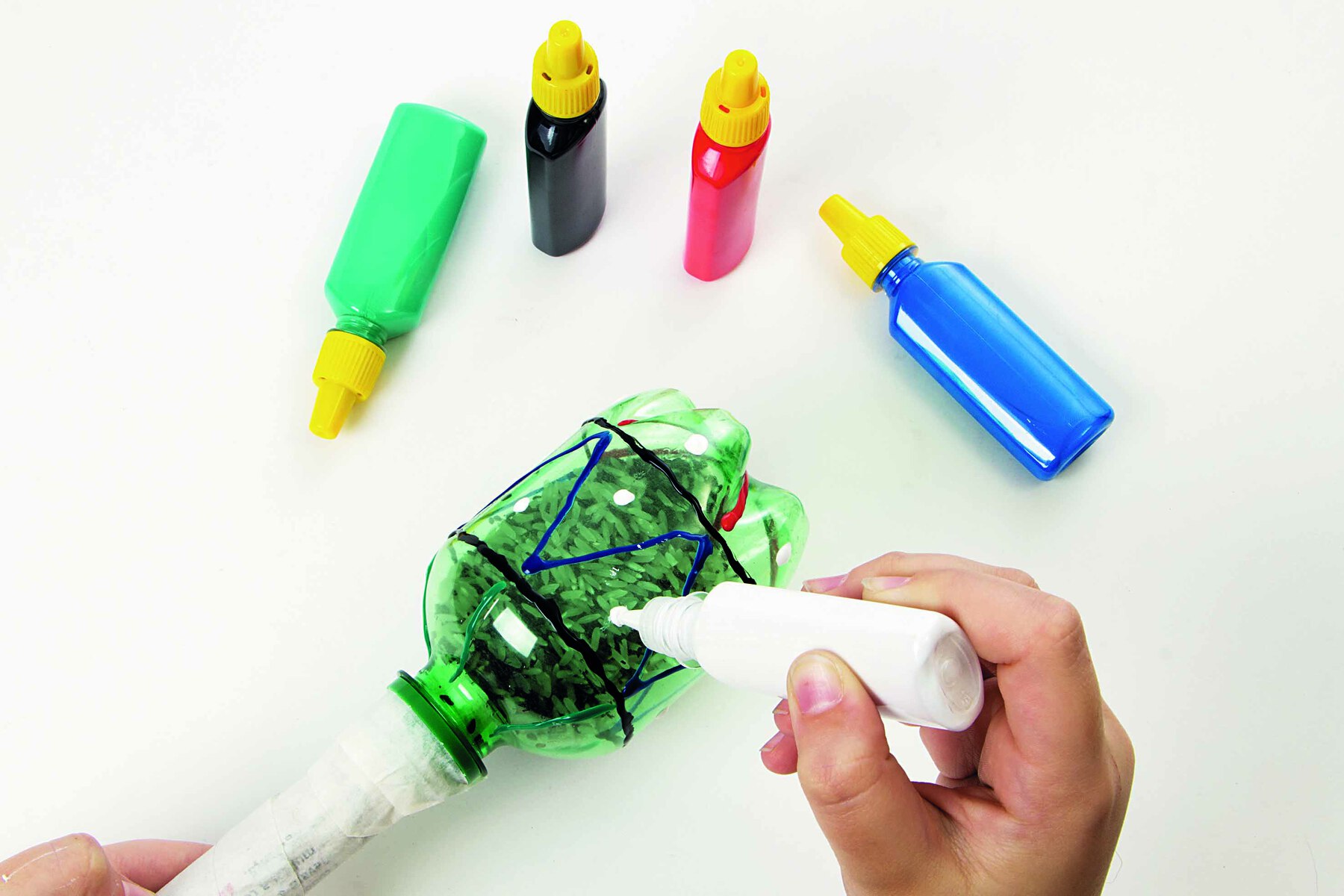 Fotografia. Destaque de mãos aplicando tinta colorida em uma garrafa PET, formando desenhos geométricos. Acima, tubos de tinta colorida.
