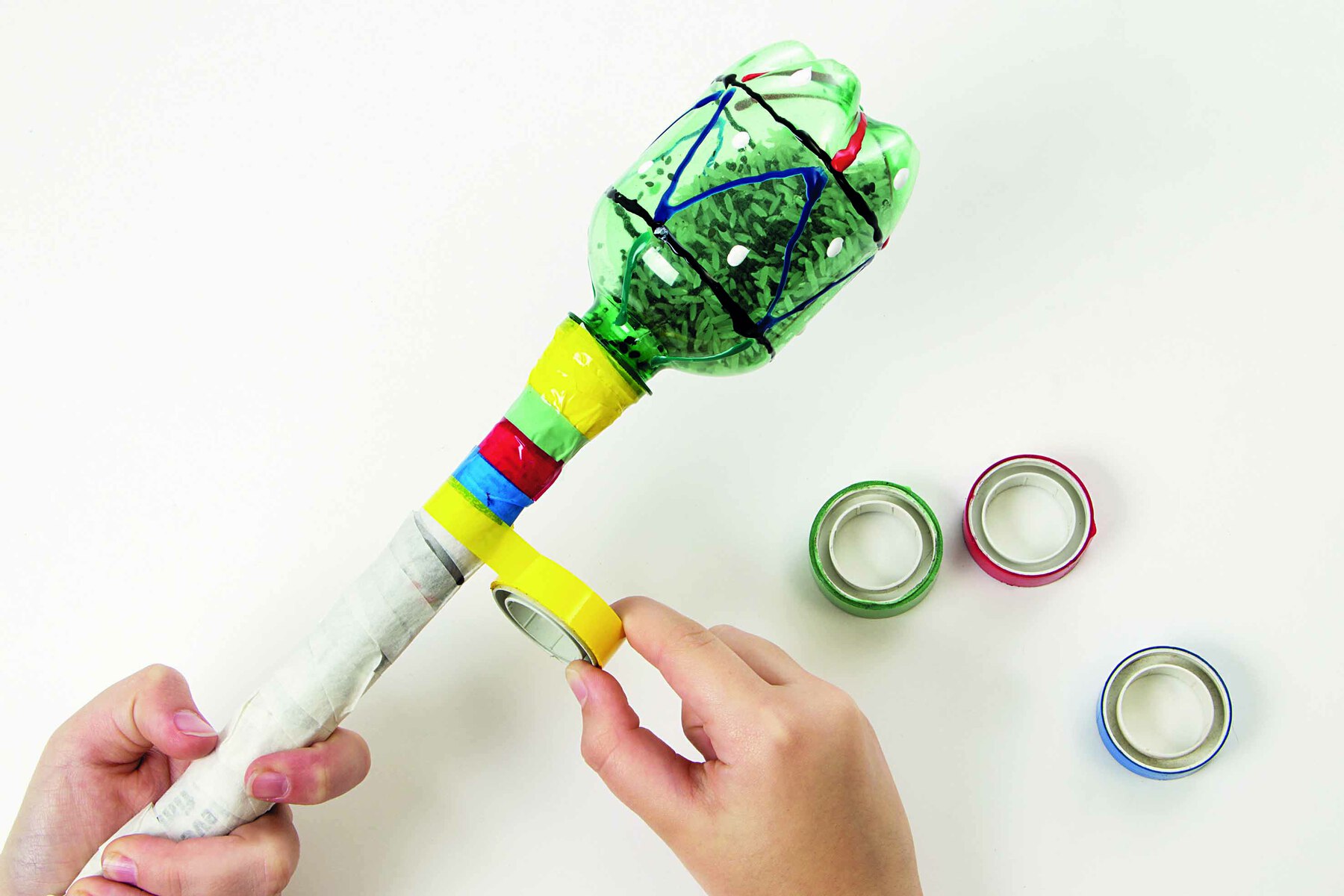 Fotografia. Destaque de mãos aplicando fitas adesivas coloridas na haste do instrumento musical construído com a garrafa PET.
