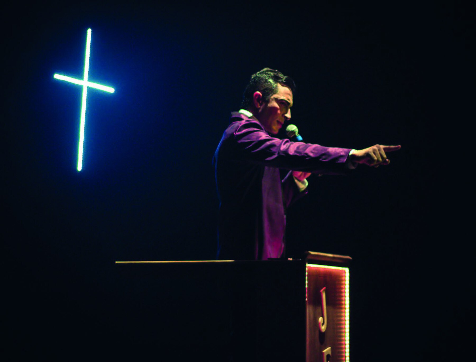 Fotografia. Um homem de cabelos pretos curtos e vestindo um  casaco roxo segura um microfone, enquanto aponta com o indicador direito para a frente, posicionado sobre um palanque. Atrás dele,  sobre fundo escuro,  há uma cruz iluminada.