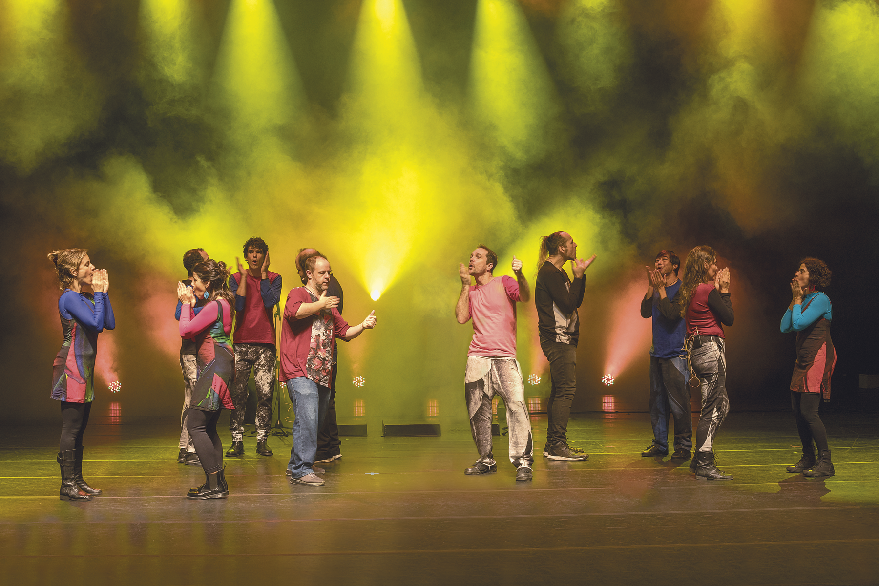 Fotografia. Em um palco iluminado com fachos de luz amarela e rosa, com fundo enevoado, homens e mulheres espalhados vestindo roupas coloridas estão com as mãos em frente à boca realizando movimentos.